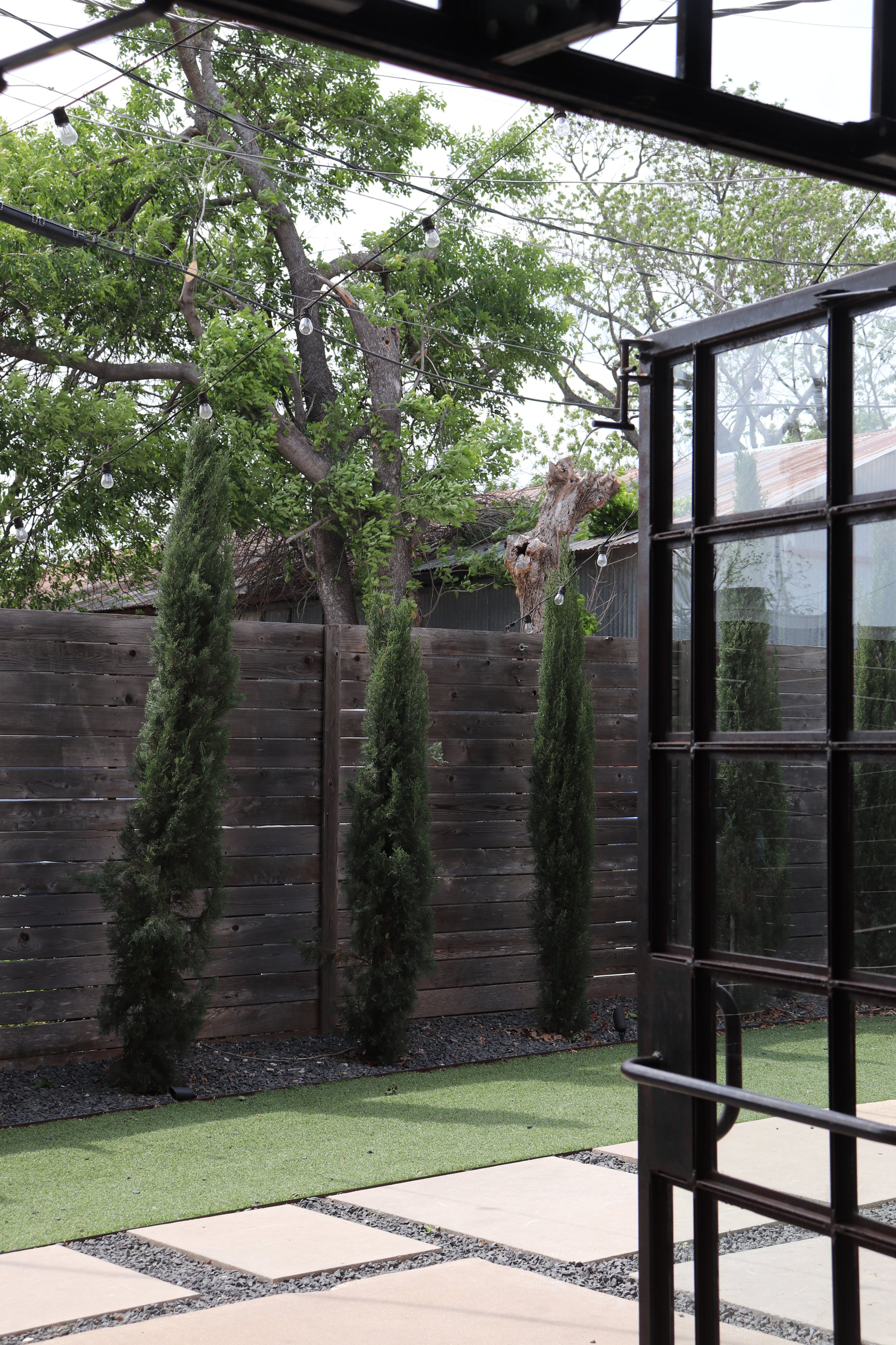 View of a backyard garden with a wooden fence, tall cypress trees, a large leafy tree, string lights, and a glass door frame.
