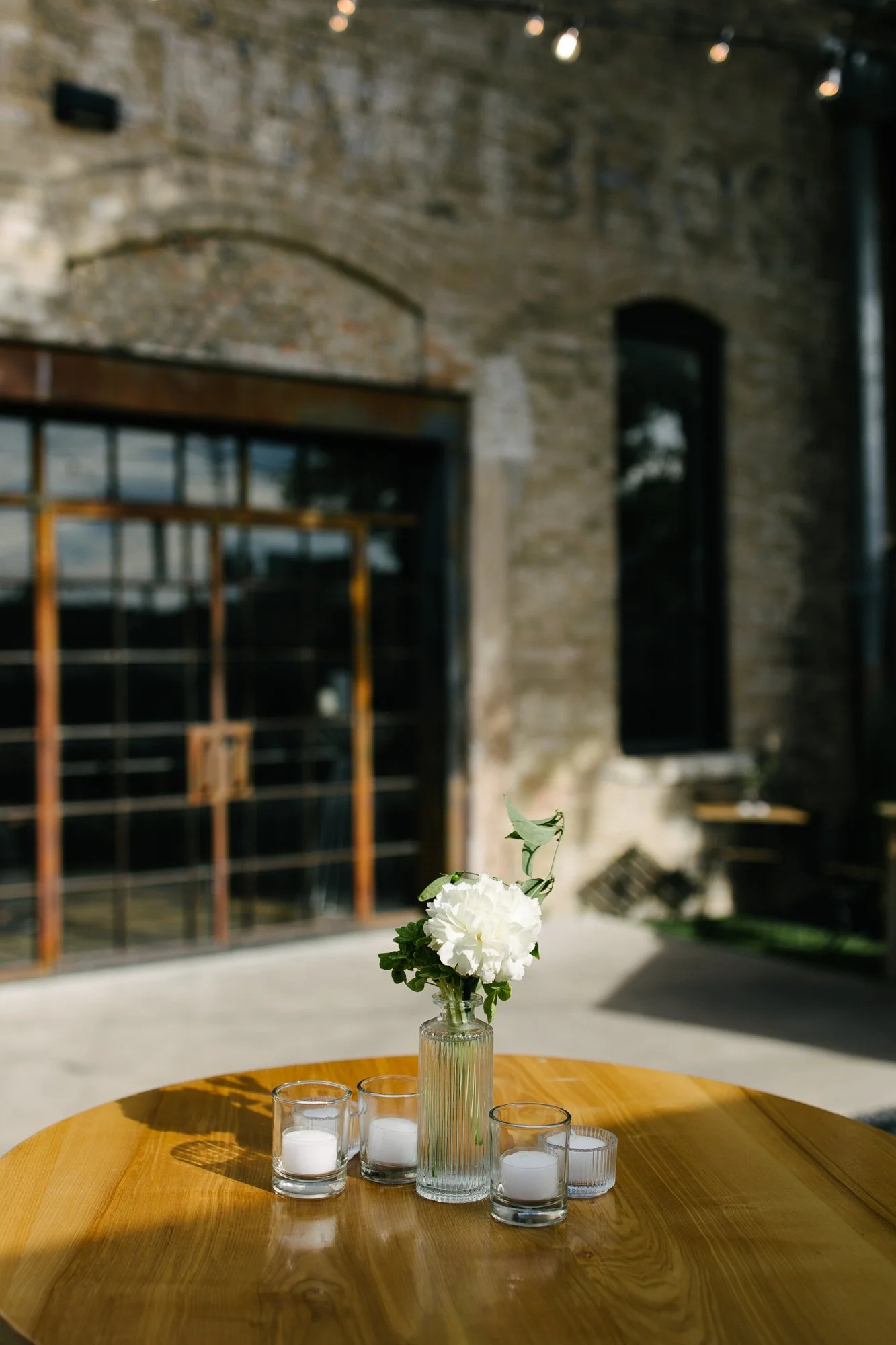 A wooden table with a glass vase holding white flowers and green leaves, surrounded by five small glass candle holders with white candles, set outdoors against a brick building with windows and a large glass door.