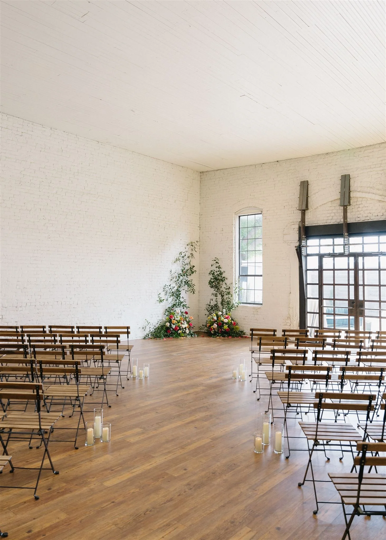 Empty room set up for wedding ceremony with wooden chairs arranged in rows and floral arrangements at the front, with candles on the floor leading to the altar.