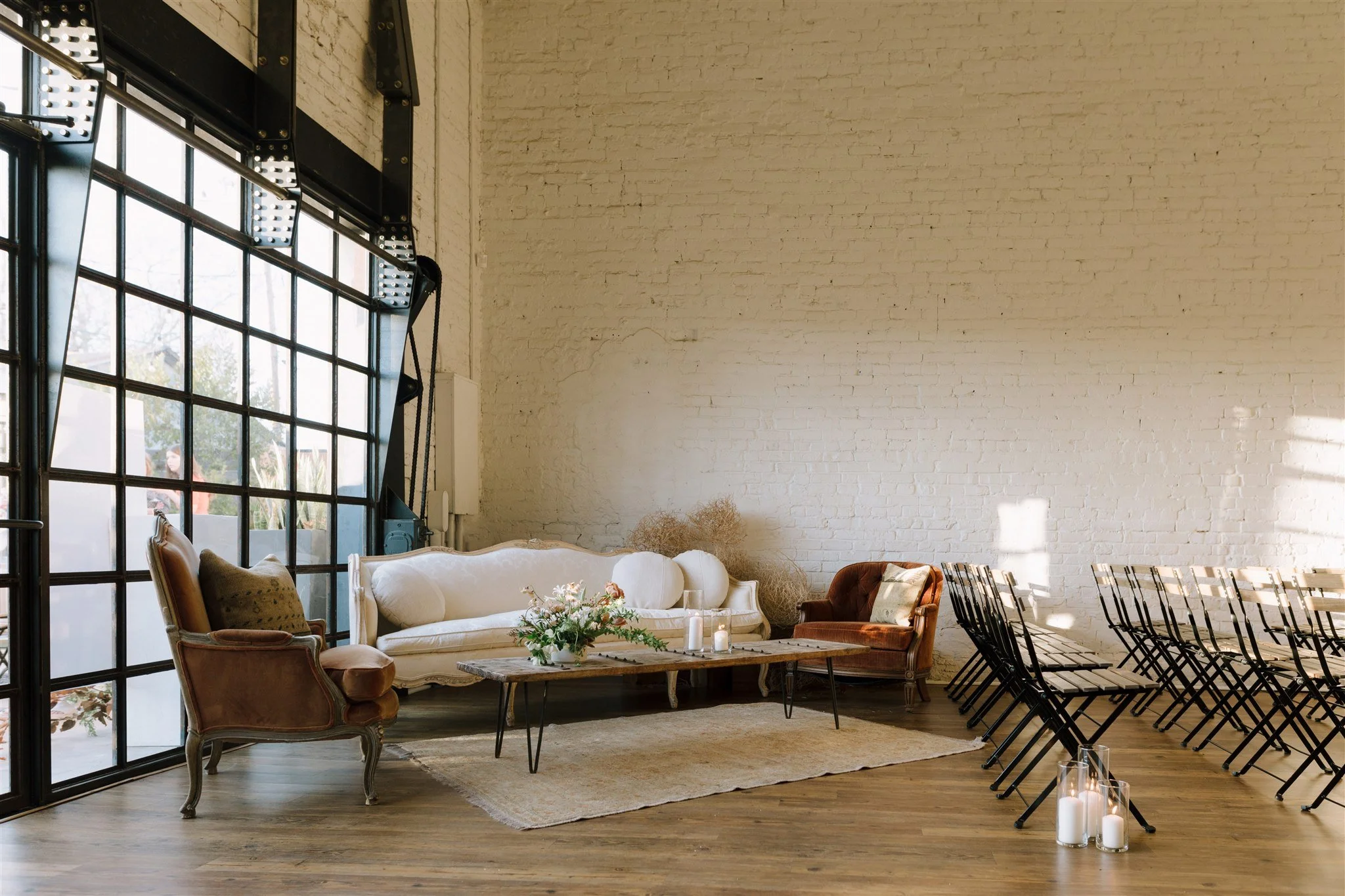 An interior space with vintage furniture arranged for a gathering, including chairs, a couch, a coffee table with flowers, candles, and a white brick wall with sunlight streaming through a large window.