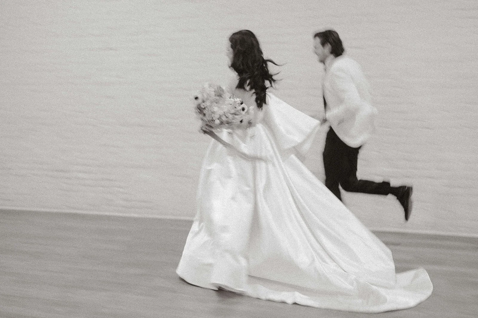 A bride in a wedding dress holding a bouquet runs on a beach with a groom in a white jacket jumping behind her.