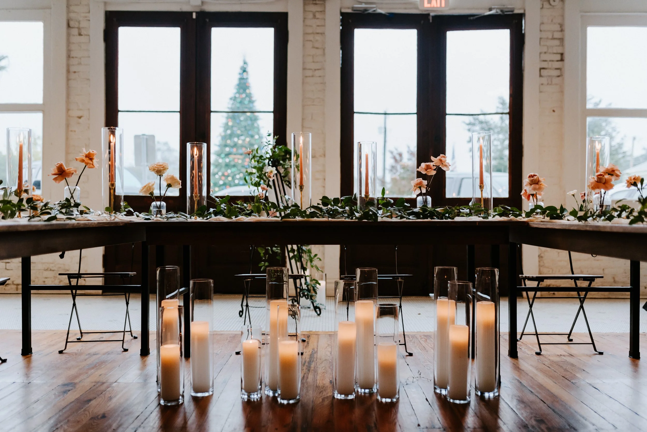 Elegant indoor holiday setting with a long table decorated with pink flowers, tall candles in glass holders, and greenery, with large windows showing a Christmas tree decorated with lights outside.