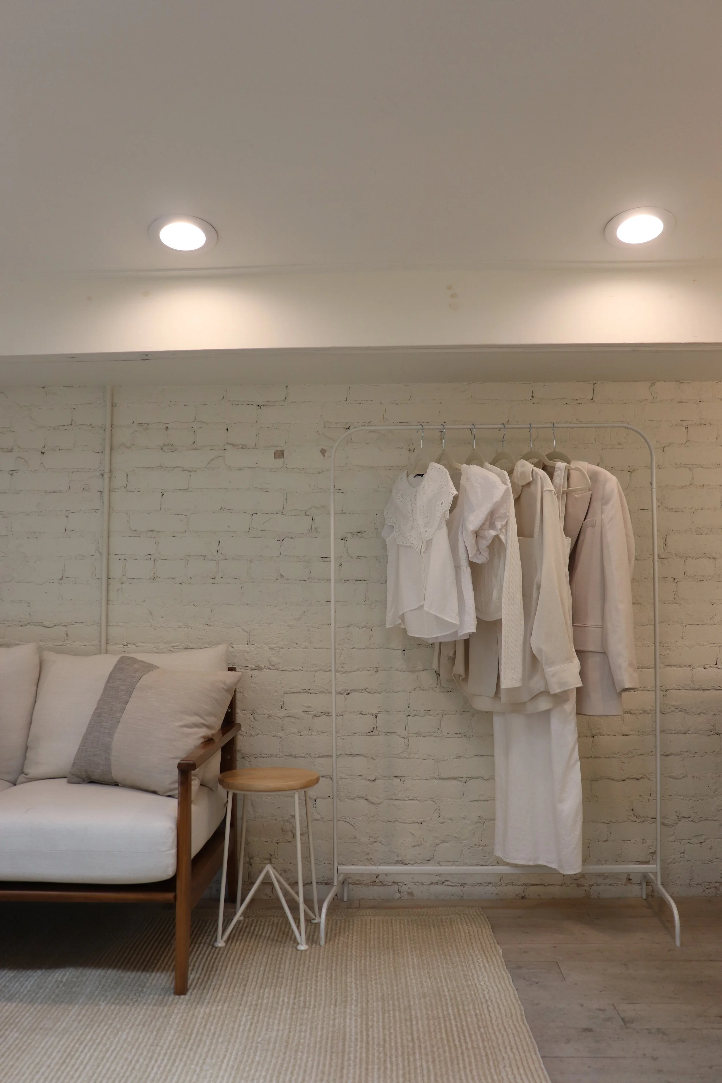 A cozy room with a white brick wall, beige couch with pillows, a small wooden side table, and a white clothing rack with white clothes hanging on it.