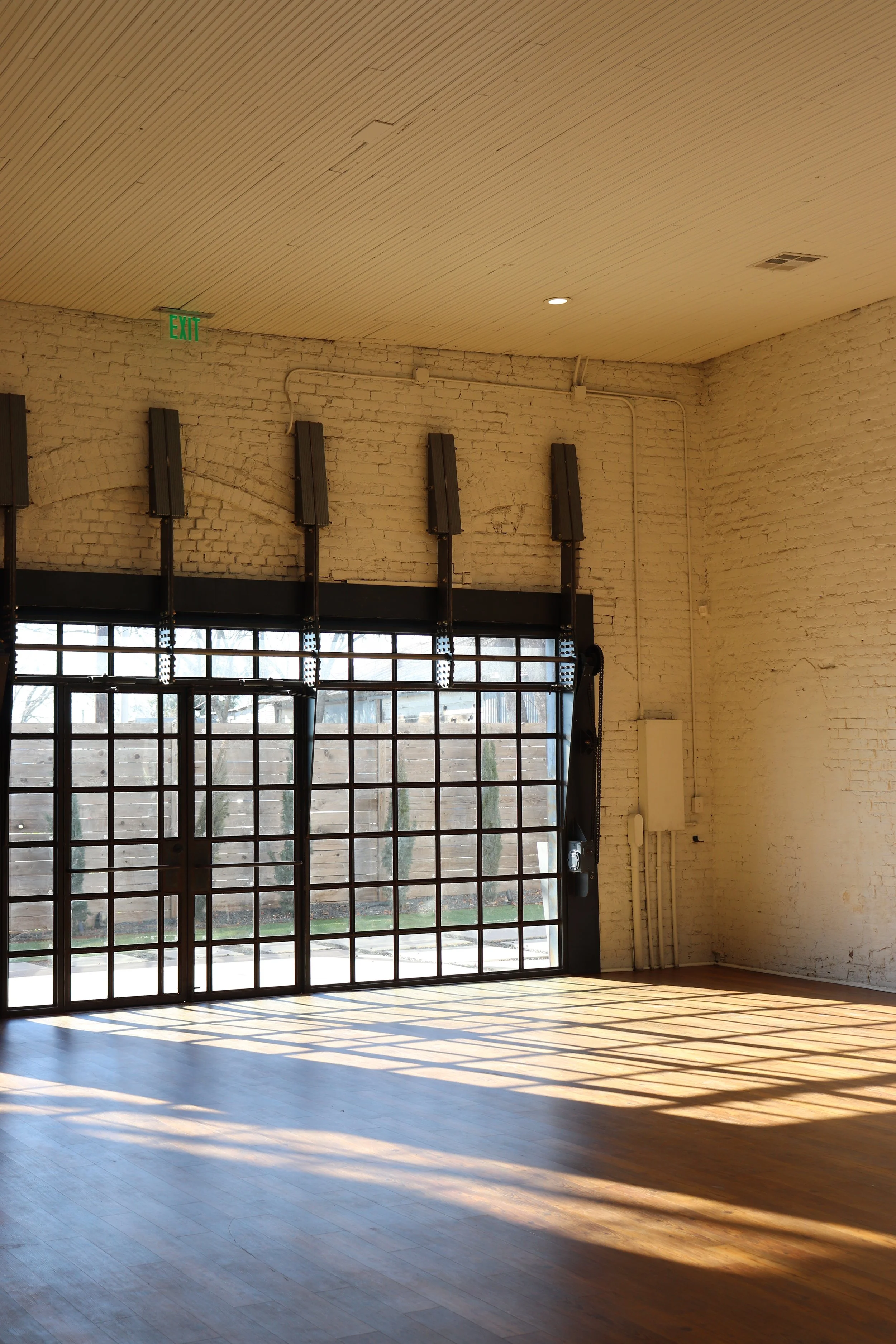 Empty room with large glass doors casting shadows on wooden floor, white brick walls, ceiling lights, and an exit sign.