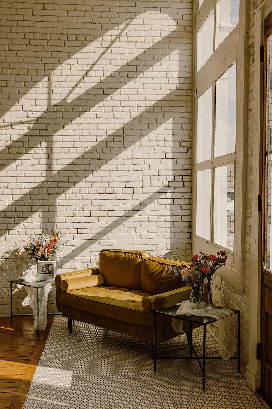 A cozy living room corner with a yellow velvet loveseat, two black metal side tables with flower arrangements, and natural sunlight streaming through large windows casting shadows on a white brick wall.