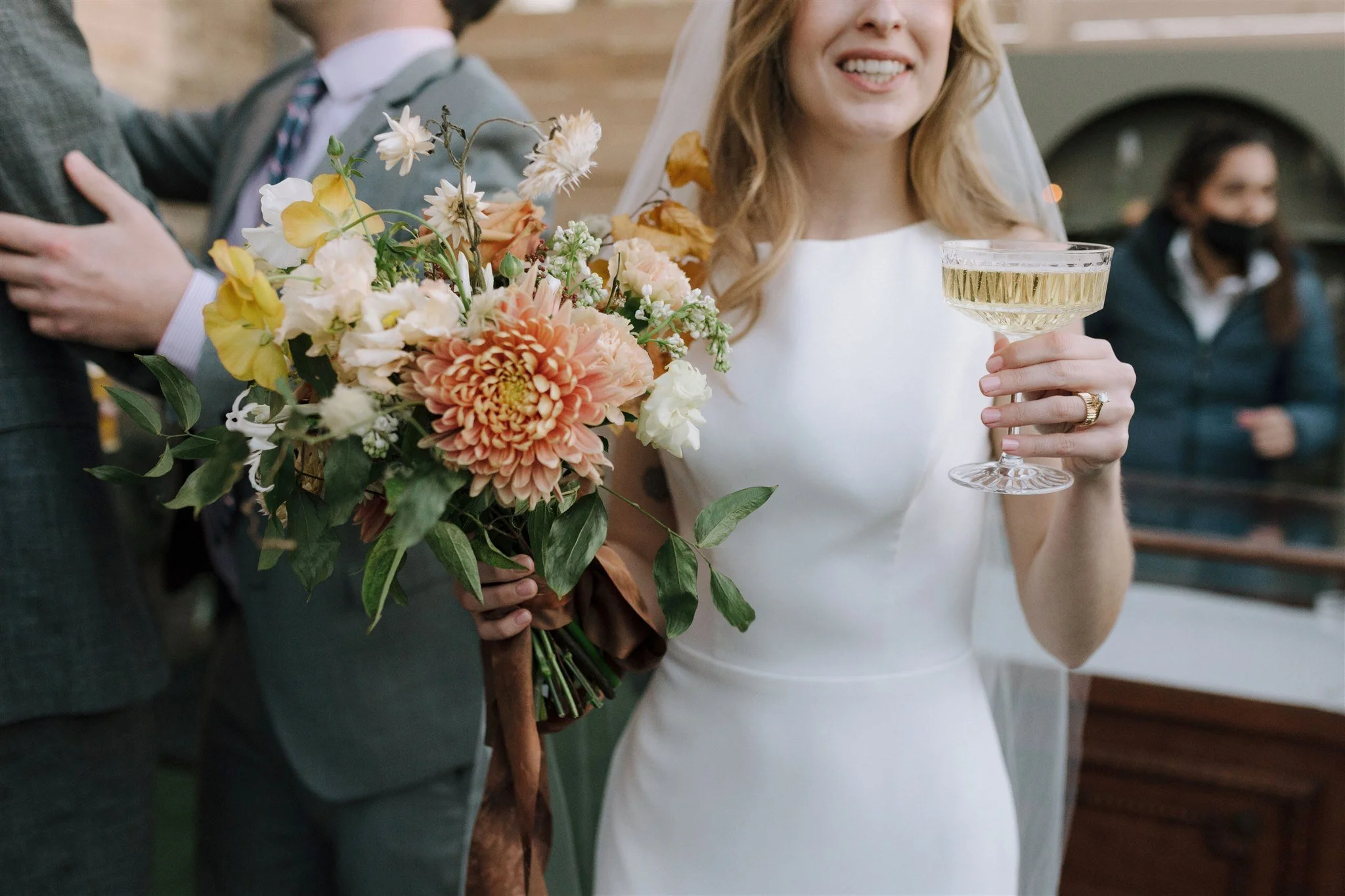 Bride in white dress holding a bouquet of flowers and a champagne flute at a wedding reception.