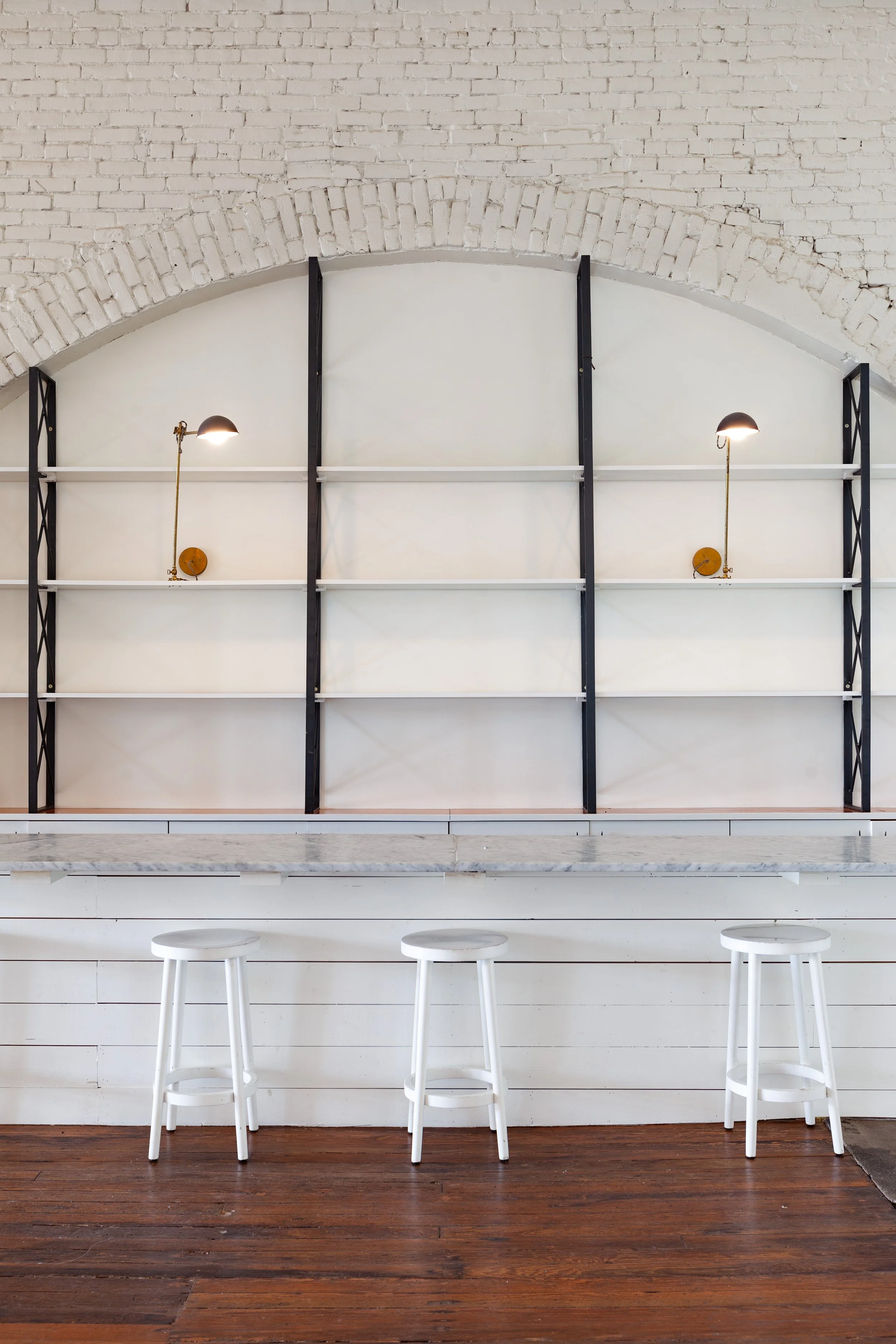 Empty white shelves with black supports on a white brick wall, illuminated by two wall-mounted lamps, above a white marble countertop and three white bar stools on a wooden floor.