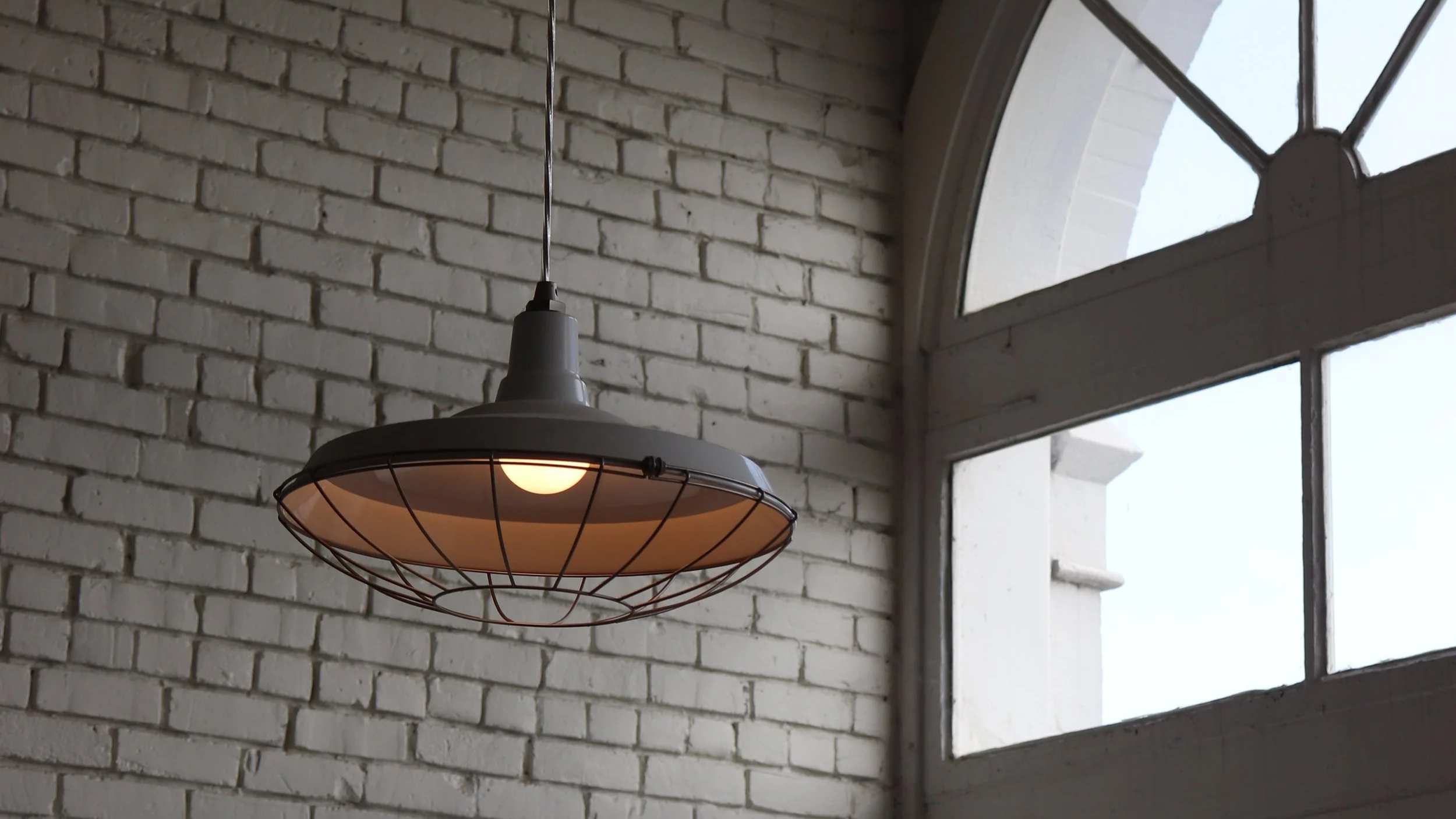 An industrial-style hanging ceiling lamp with a metal wire cage surrounding a round light bulb, hanging near a large arched window with multiple panes, against a white brick wall.