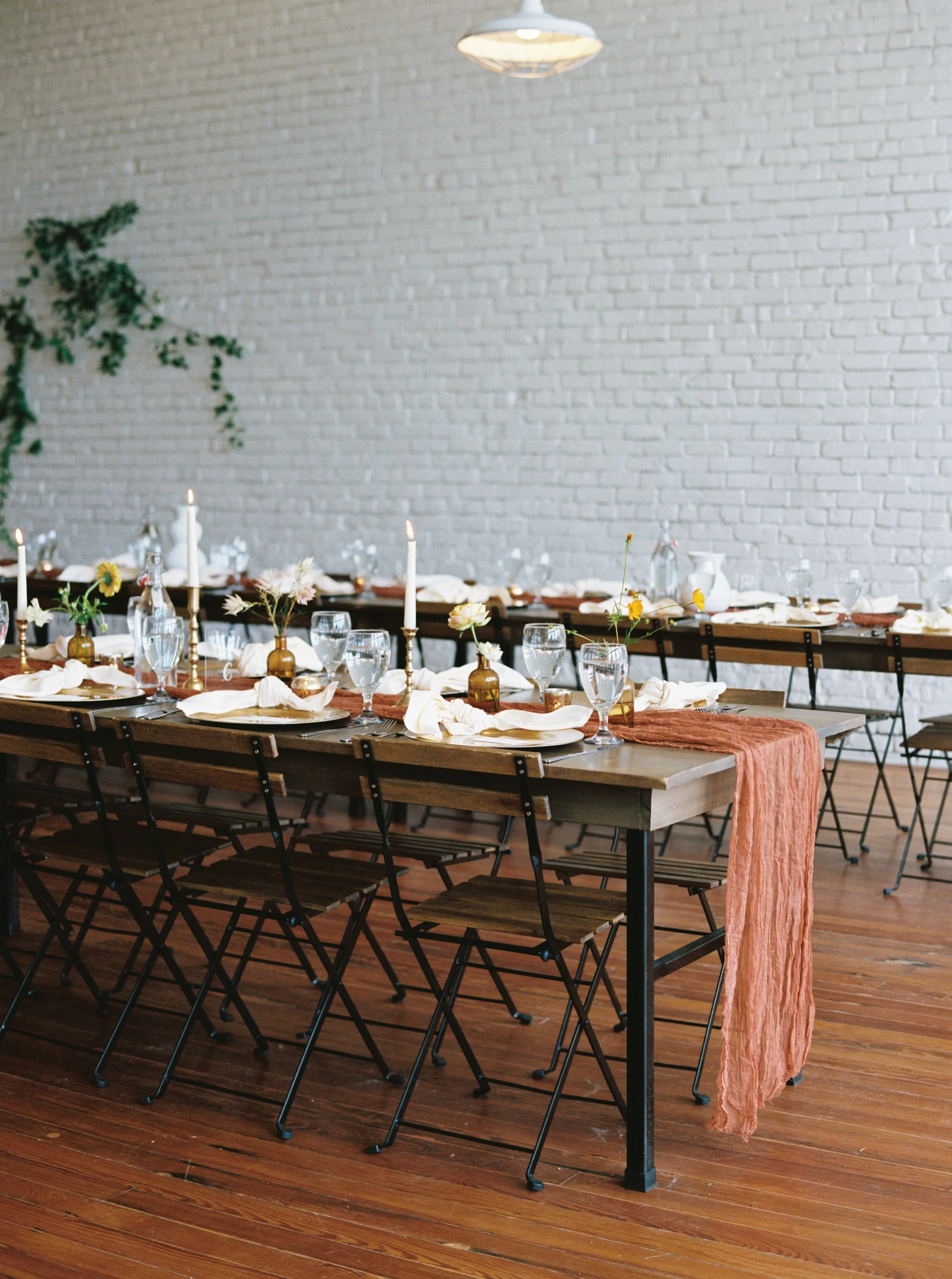 Decorated dining table set with white napkins, glassware, candles, and small flower arrangements in bottles, in a bright room with a white brick wall and wooden floor.