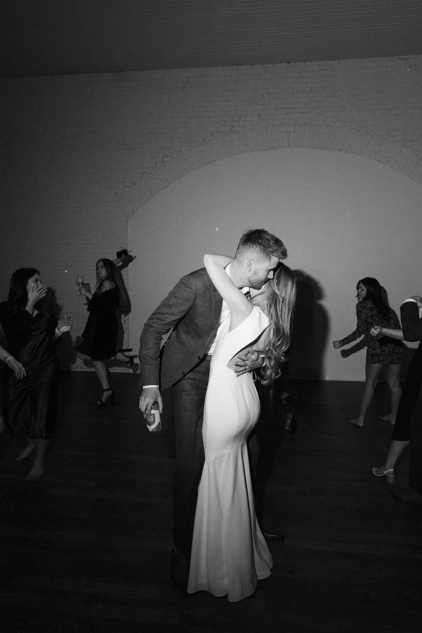 A couple dancing closely at a party, with the man dressed in a suit and the woman in a long white dress, surrounded by other partygoers in a dimly lit room.
