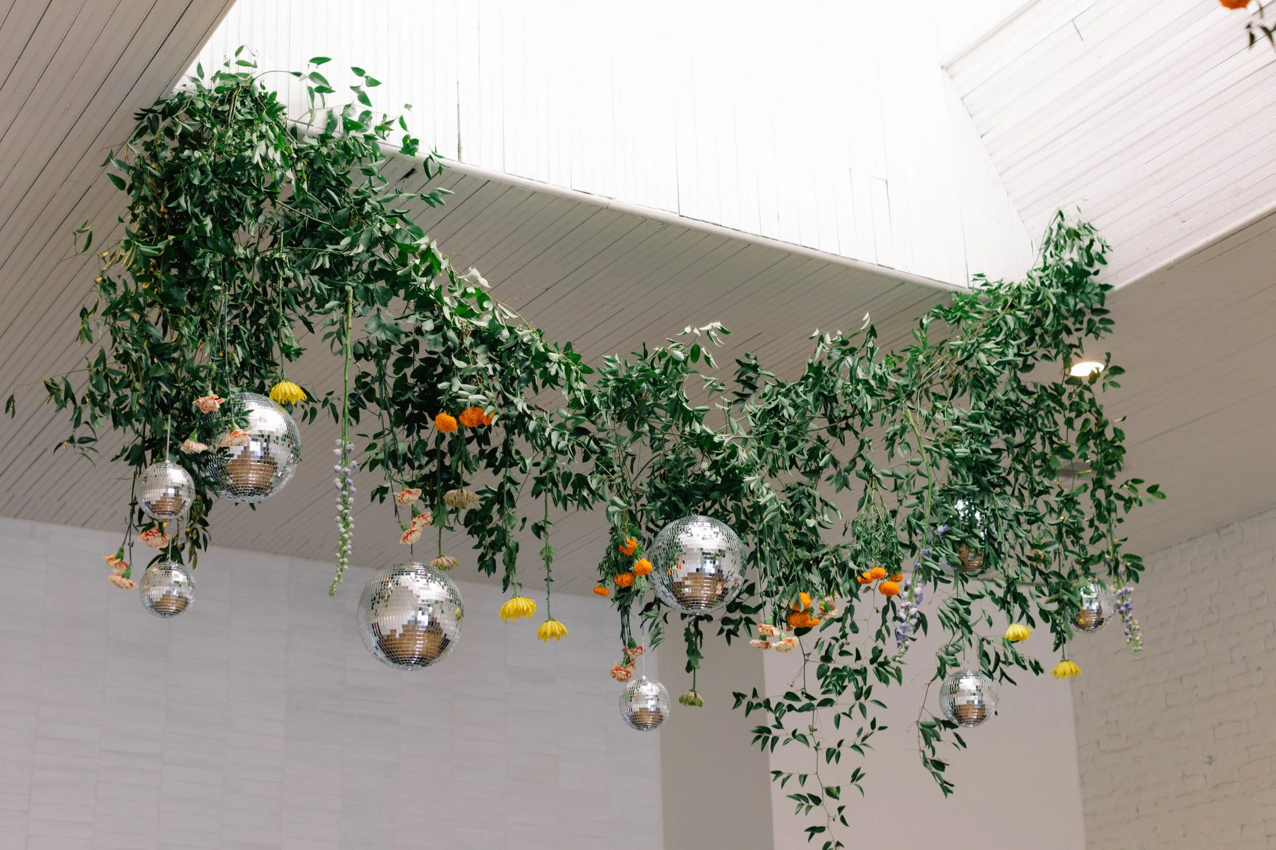 Decorative greenery with disco balls and colorful flowers hanging from the ceiling in a white room.