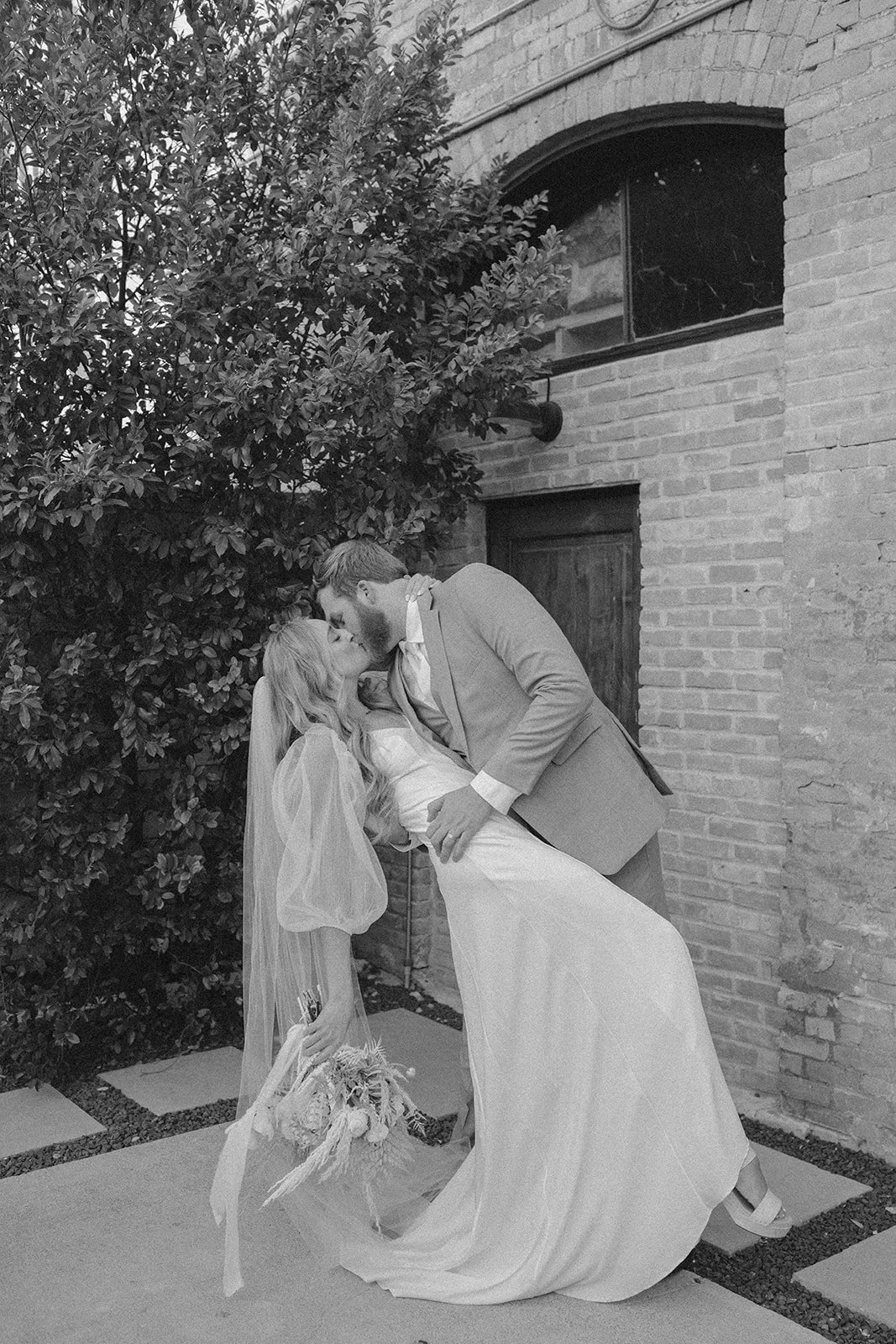 A bride and groom are sharing a kiss outside a brick building, with the groom dipping the bride. The bride is holding a bouquet of flowers, and there is a tree and a window in the background.