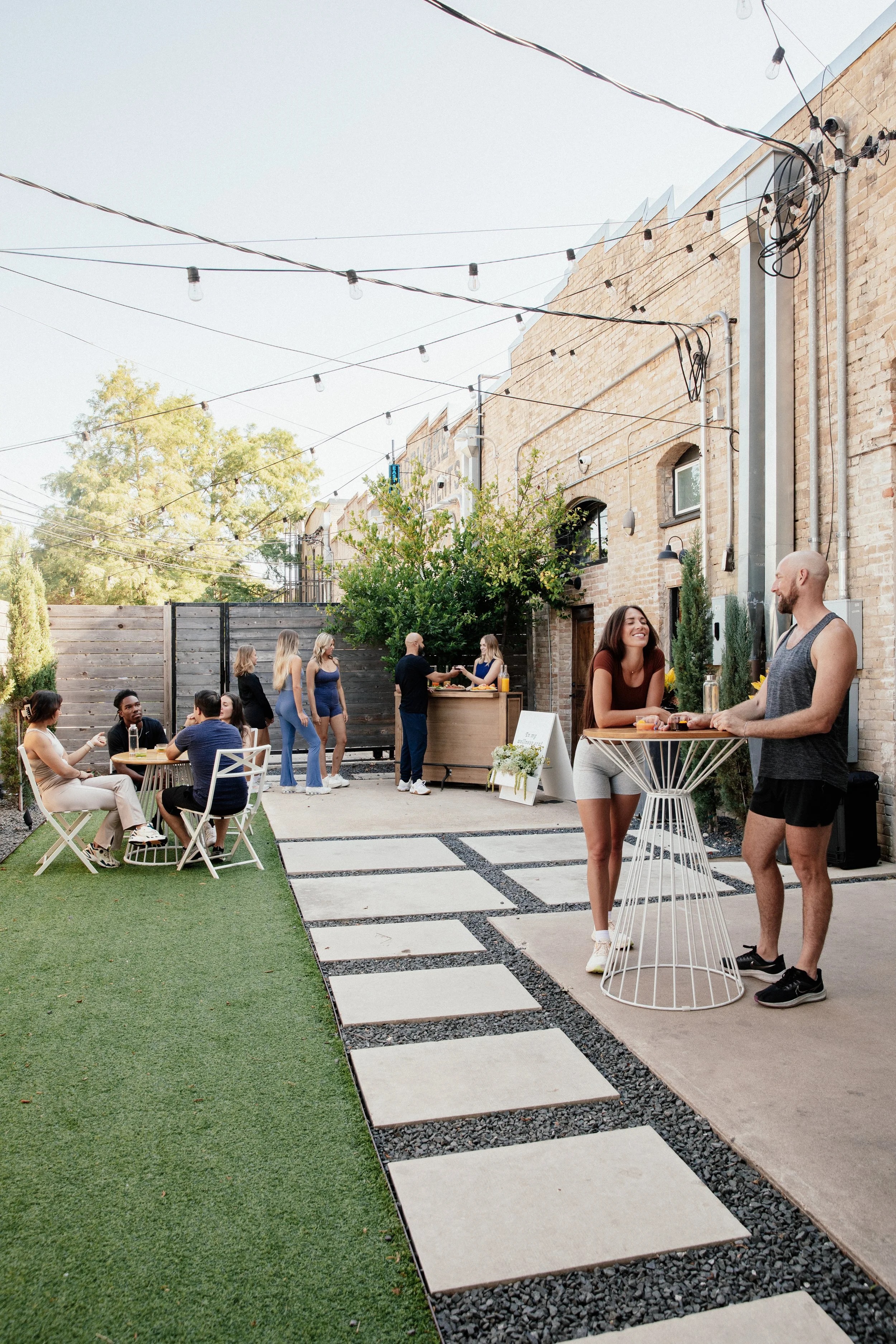 People at an outdoor gathering with string lights, a wooden fence, and brick buildings in the background. Some are sitting at small tables, others are standing and chatting, with a bar area in the background.