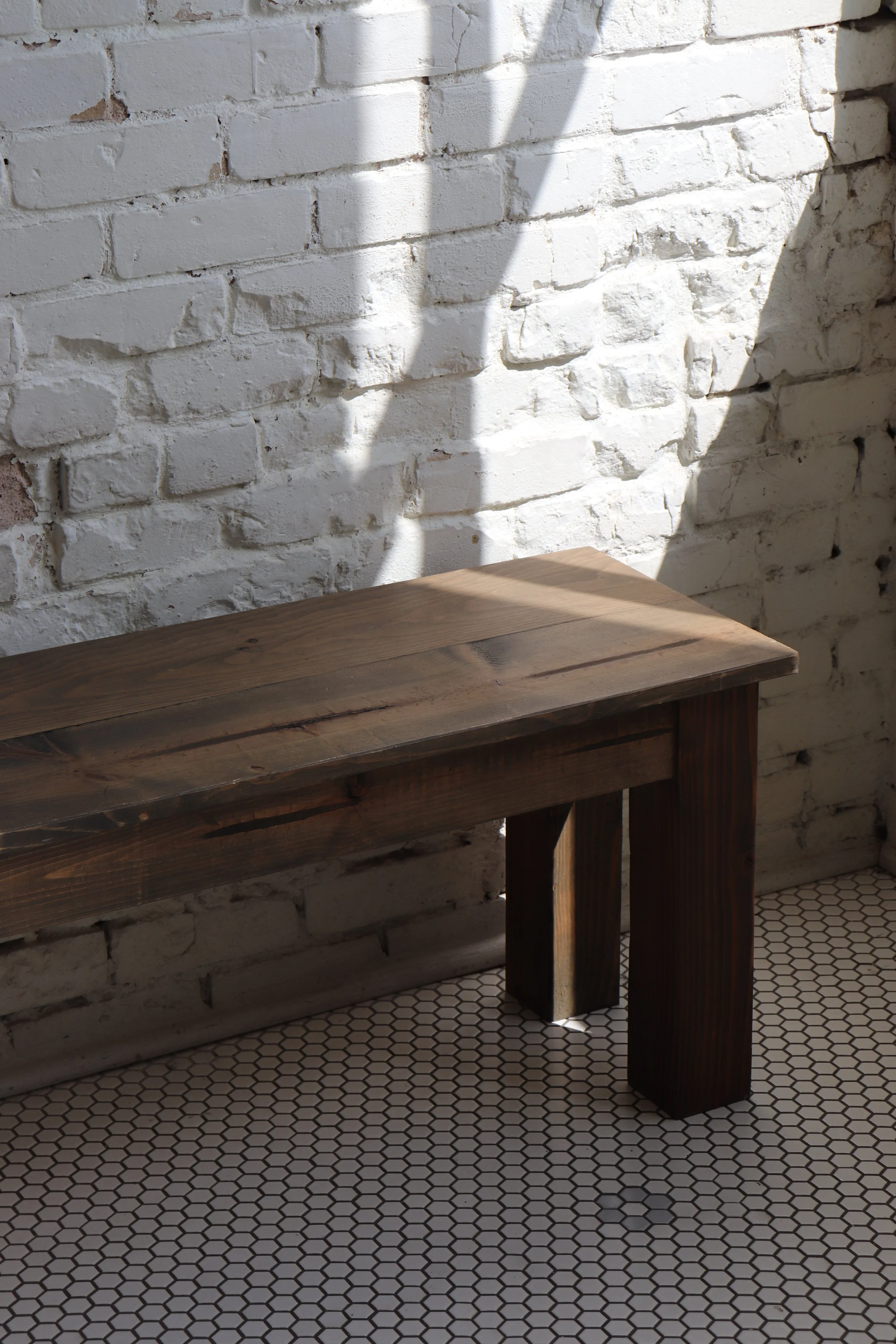 A wooden bench against a white brick wall with sunlight casting shadows on the wall and bench, and a hexagonal tile floor.