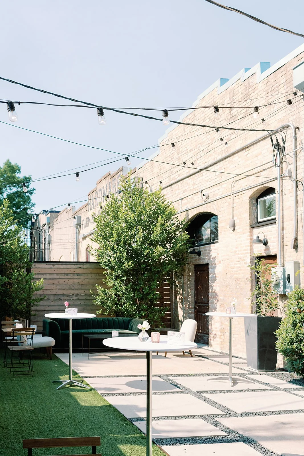 Outdoor patio with high tables, a green sofa, a white chair, and potted plants, enclosed by a wooden fence, with string lights overhead against a brick building with windows and greenery.