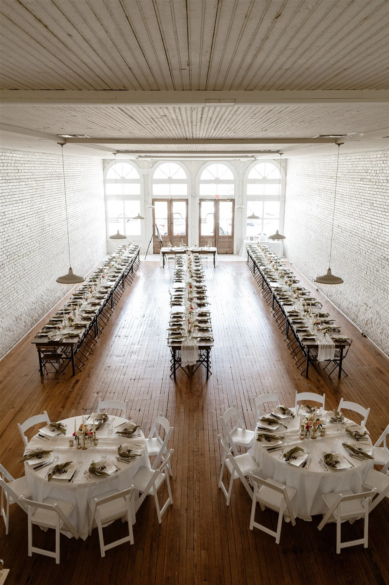 A spacious banquet hall with long tables and round tables set for a wedding or event, featuring white tablecloths, elegant place settings, and floral centerpieces, with large windows at the back allowing natural light to fill the room.
