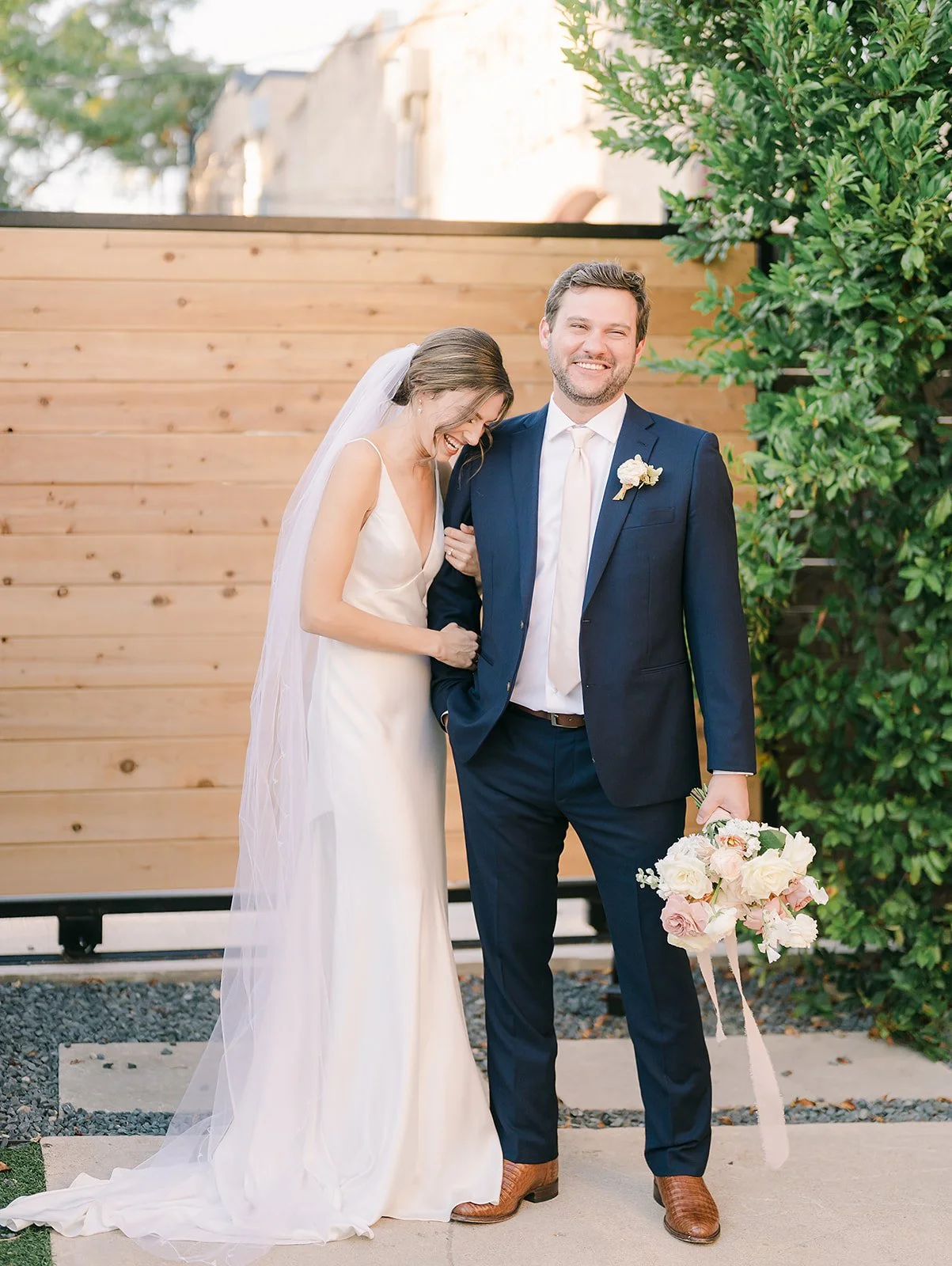A bride in a white wedding gown and veil laughing with a groom in a navy suit holding a bouquet of flowers, standing outside in front of a wooden fence and greenery.