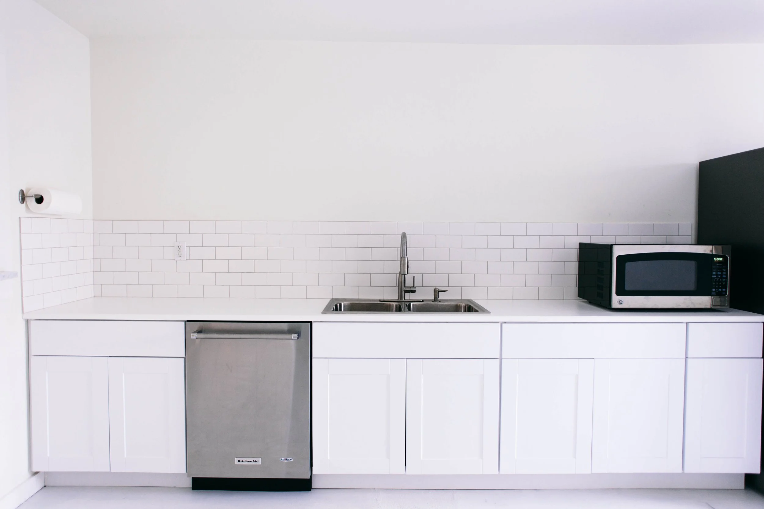 Minimalist white kitchen with white cabinets, a stainless steel dishwasher, a double sink, a microwave oven, and a partial view of a paper towel roll on a white wall with subway tile backsplash.