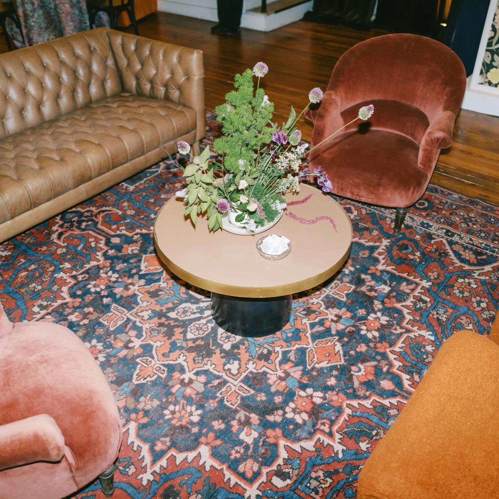 Living room with a patterned area rug, a tan tufted leather sofa, a rust-colored velvet armchair, and a round coffee table with a floral arrangement and a glass ashtray.