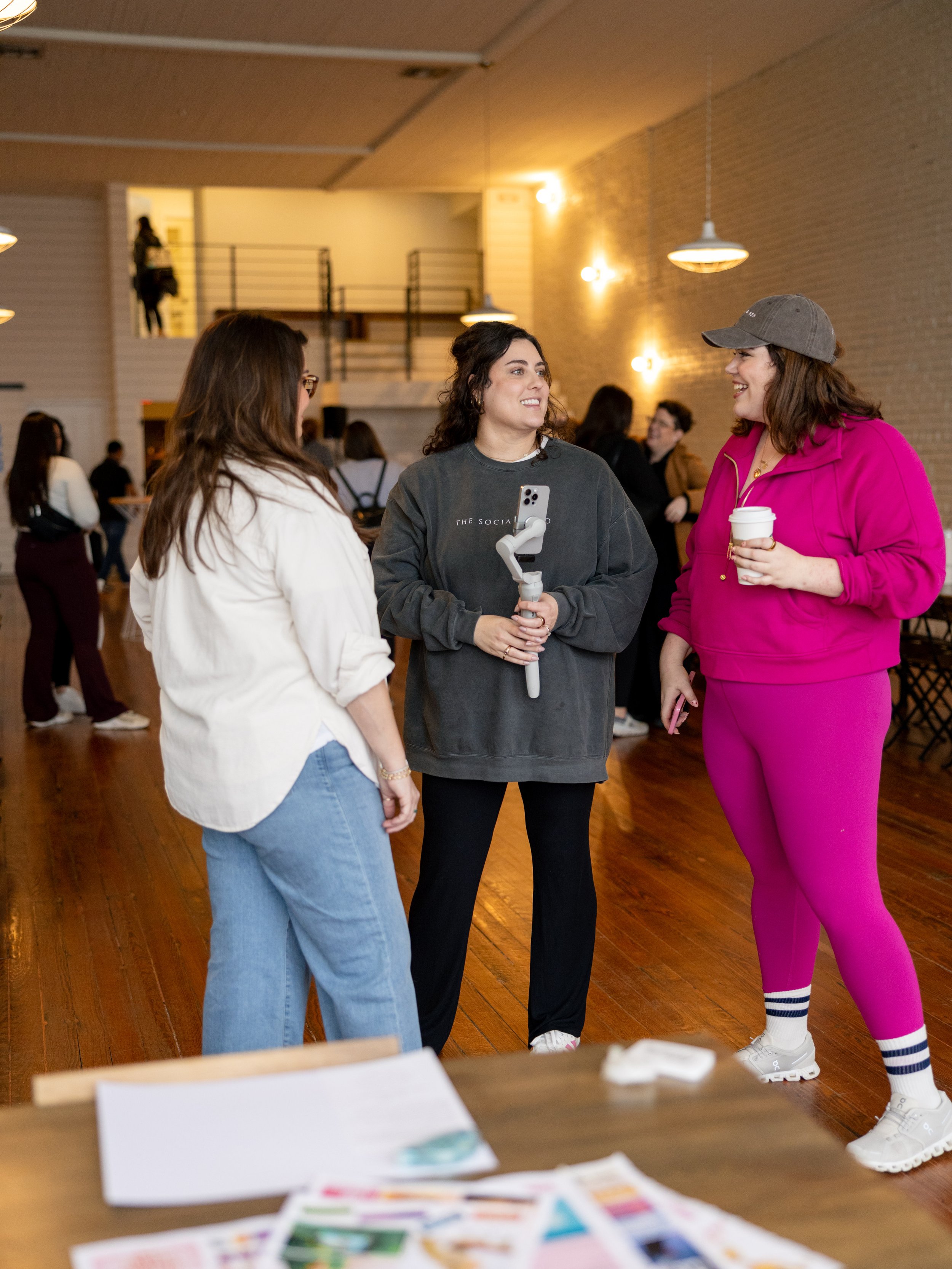 Three women are standing and talking in a social gathering inside a room with wooden floors and exposed brick walls. One woman is holding a smartphone mounted on a stabilizer, and another woman is holding a coffee cup. The setting appears lively with other people in the background.