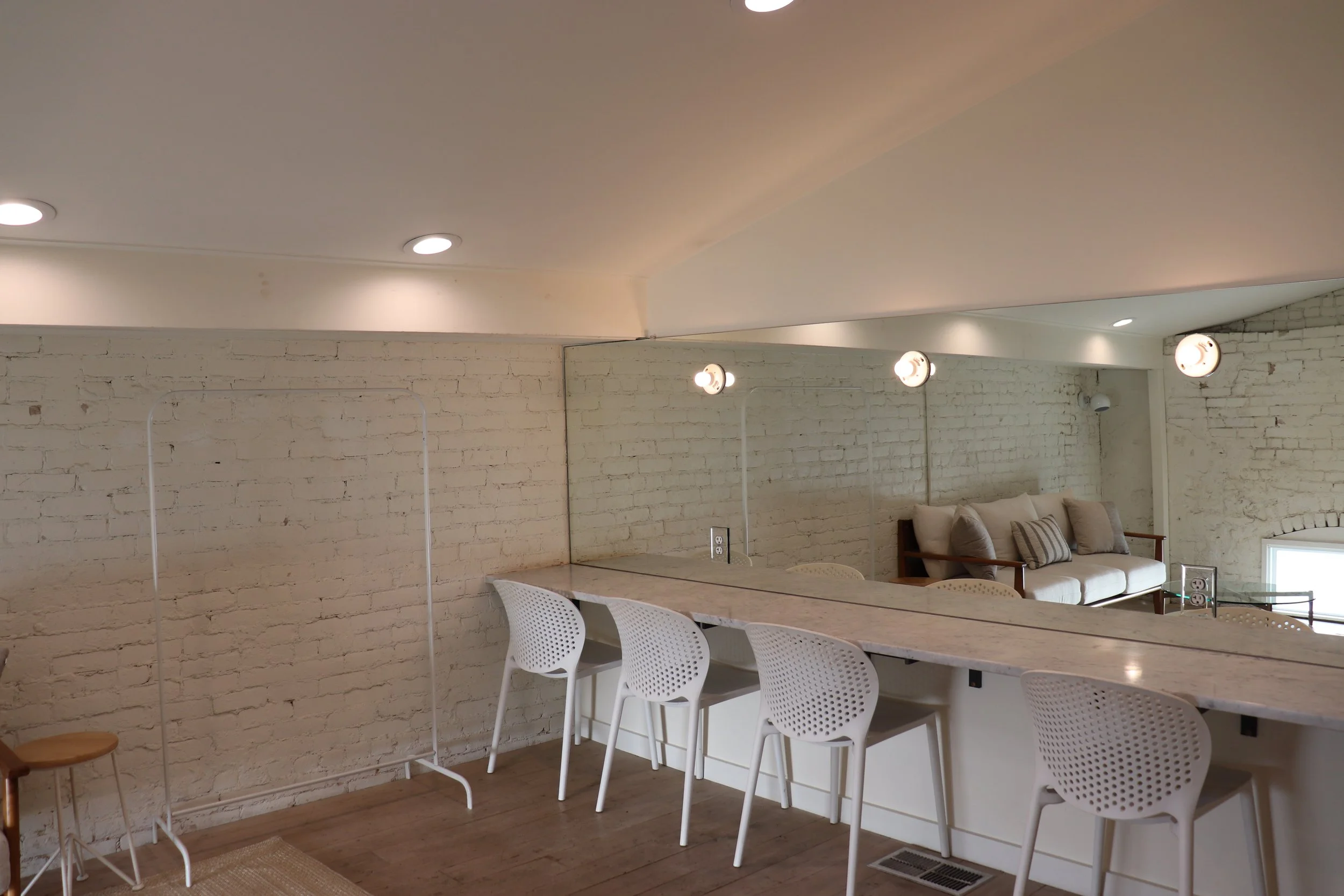 Interior of a modern room with white brick walls, a marble bar counter with white perforated chairs, a large mirror, ceiling lights, and a beige sofa with pillows.