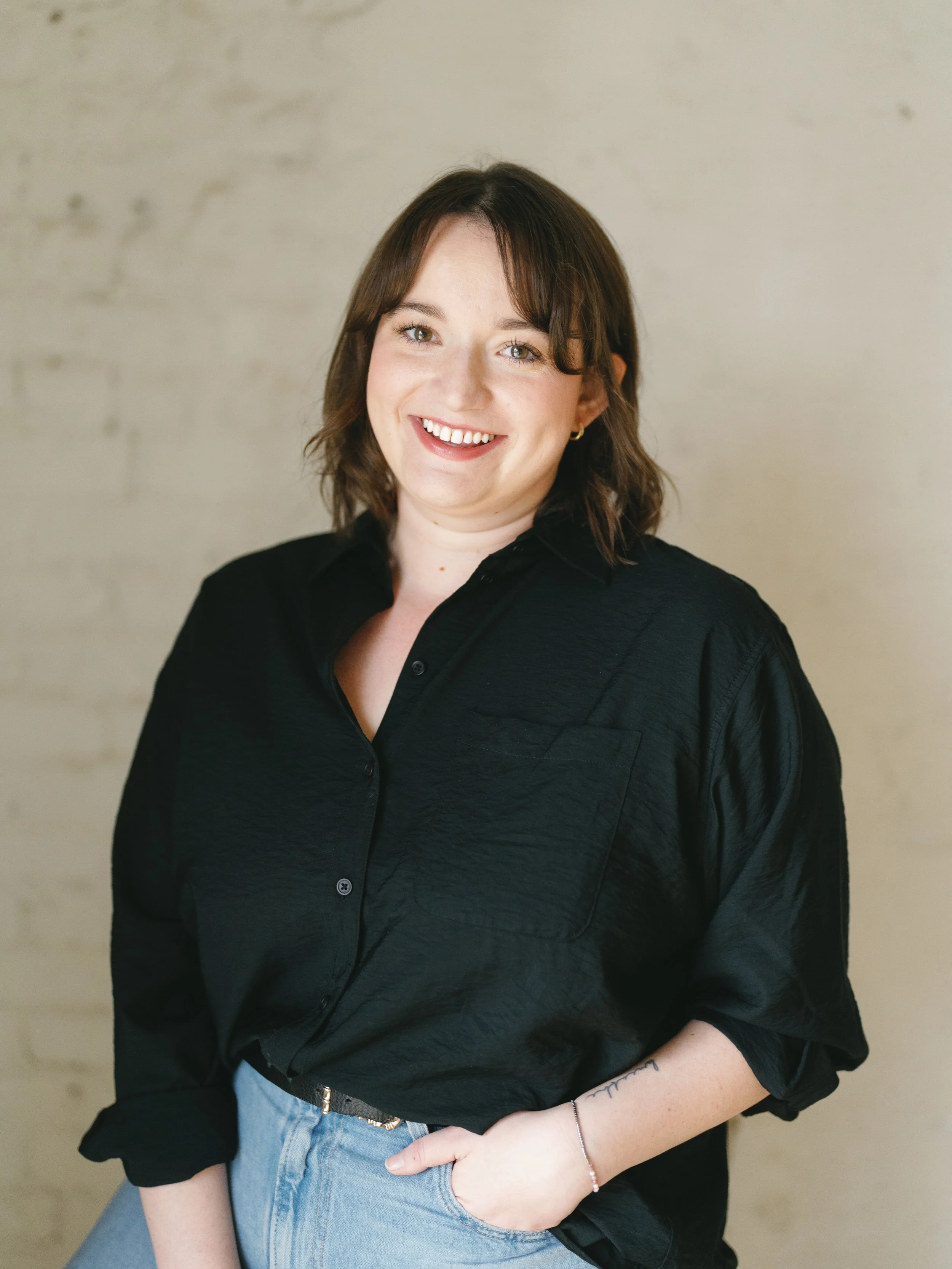 A smiling woman with shoulder-length brown hair, wearing a black button-up shirt with rolled sleeves and light blue jeans, standing against a beige wall.