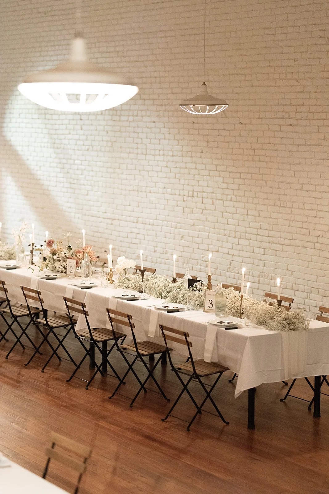 Long dining table decorated with white tablecloth, floral arrangements, candleholders, and table numbers, set against a white brick wall with hanging ceiling lights.