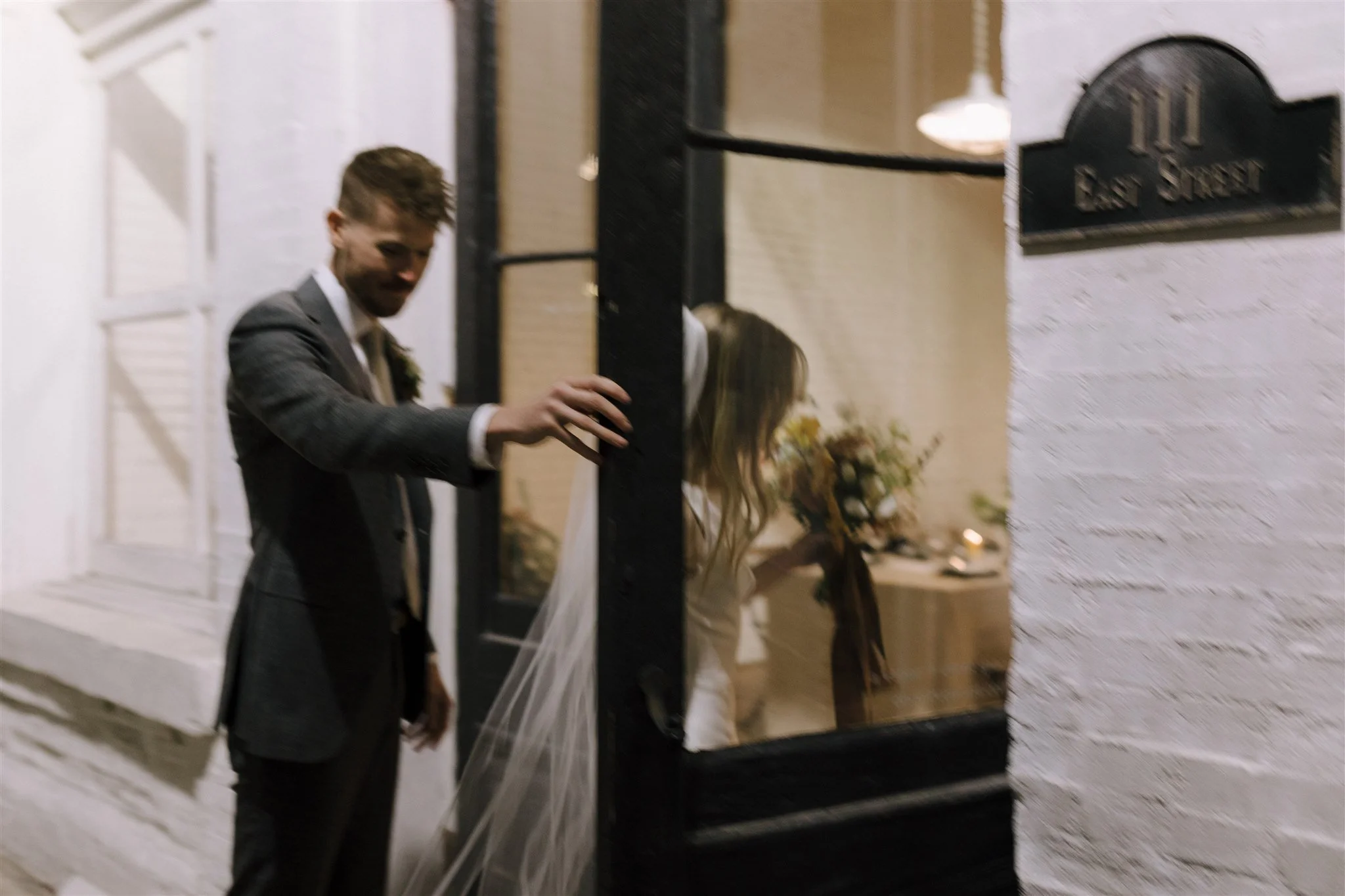 A couple dressed in wedding attire, with the groom opening a door for the bride, inside a decorated venue at night.