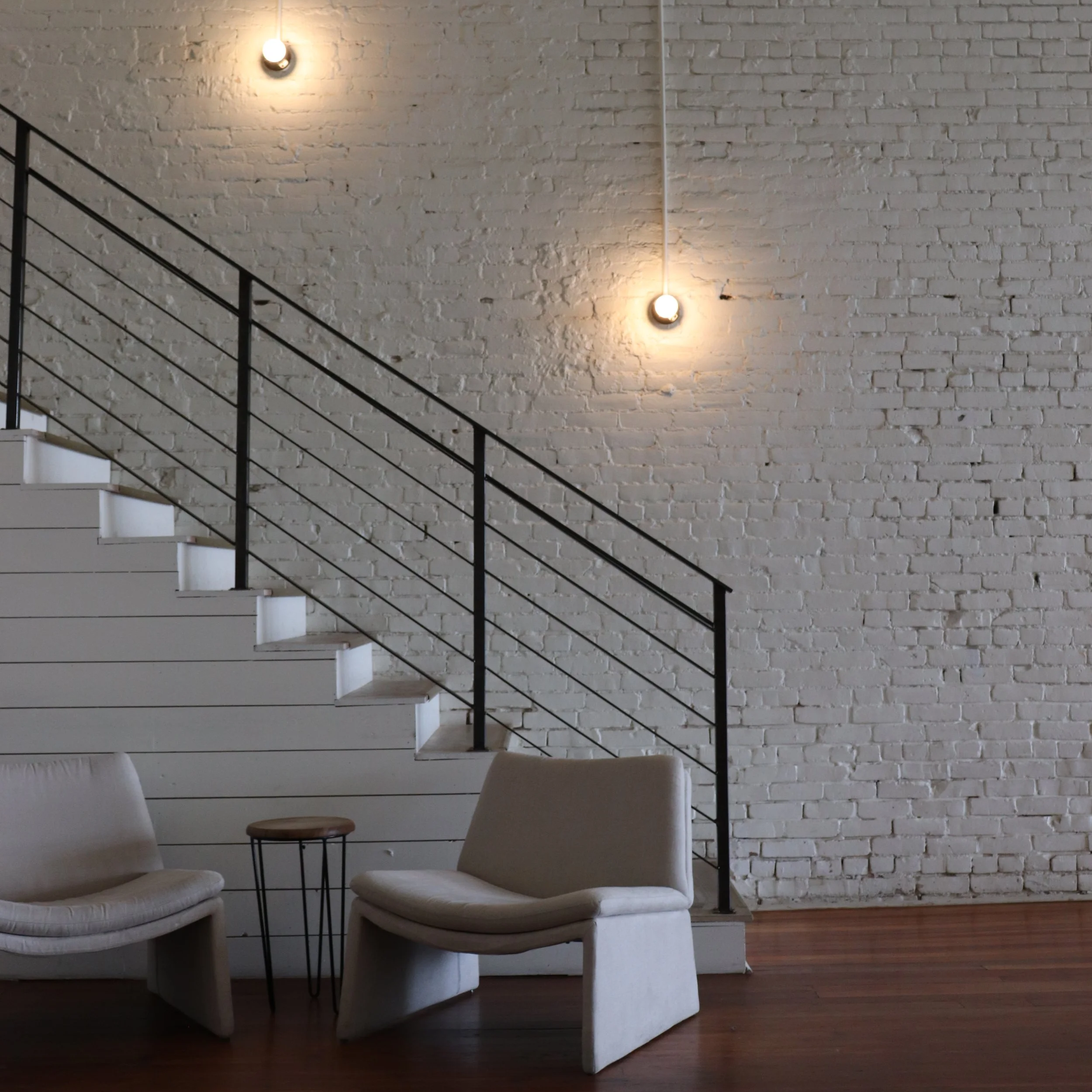 Interior of a modern space with white brick wall, black metal staircase, two beige chairs, and a small round wooden table, illuminated by two exposed light bulbs.