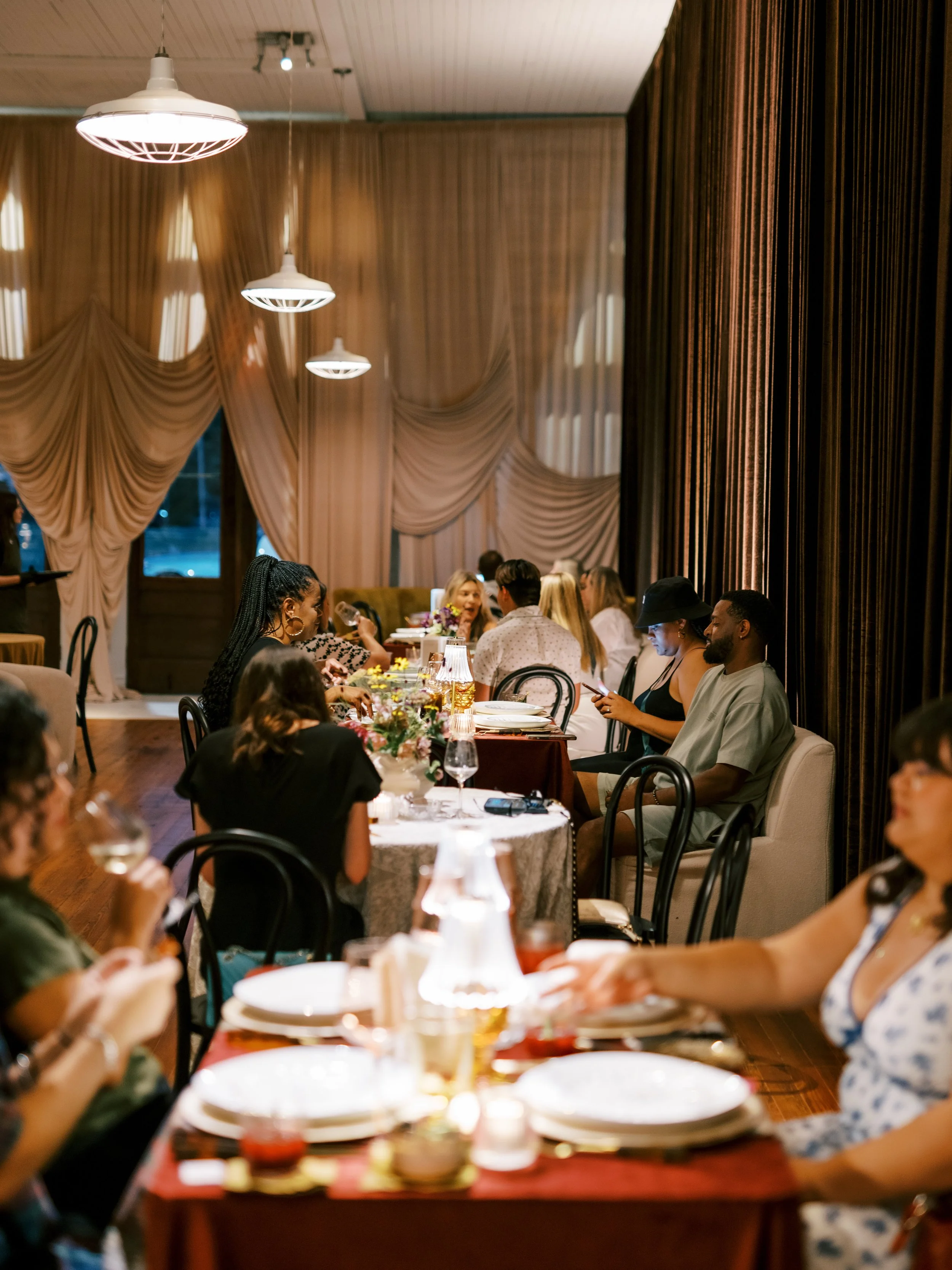 People dining at a long table in an elegant, warmly lit restaurant with draped curtains and hanging lights.