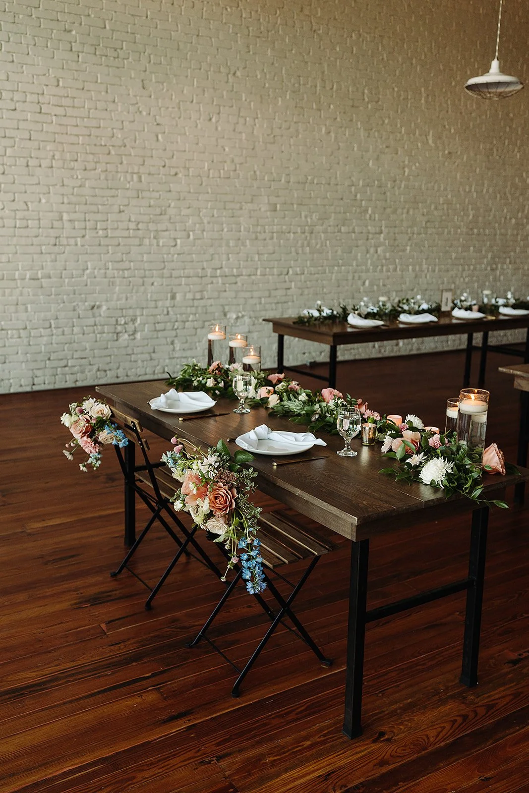 Wedding reception table decorated with pink and white flowers, candles, and white napkins, set against a white brick wall.