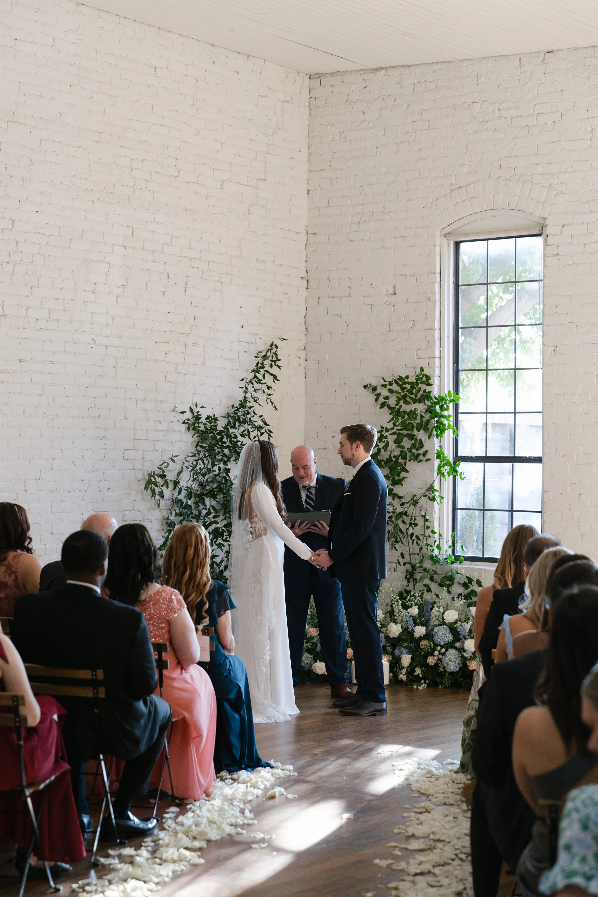 A wedding ceremony taking place indoors with a bride and groom holding hands while saying their vows, officiated by a man. Guests seated on chairs are watching, and there is floral decor and a large window in the background.