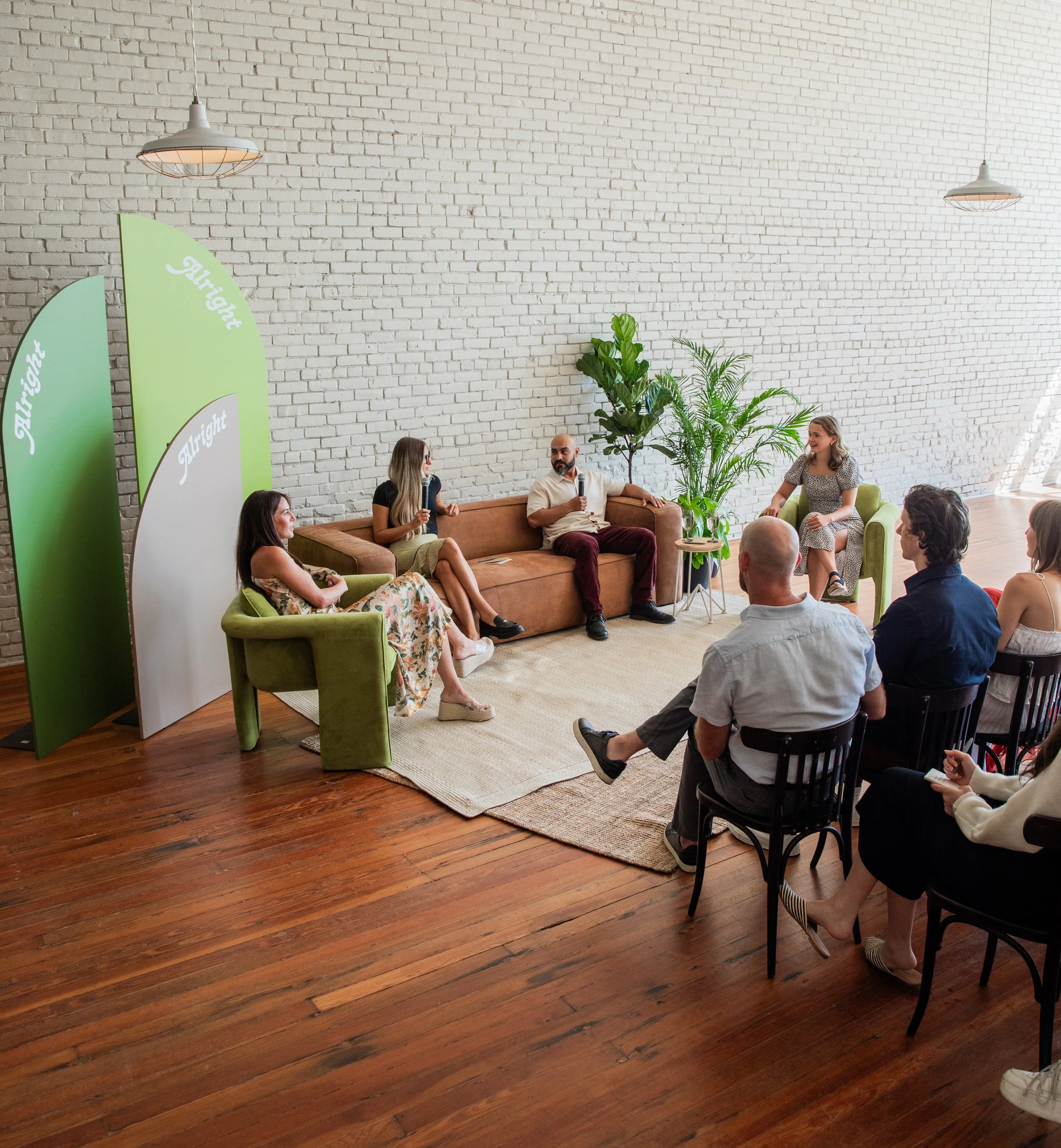 A panel discussion with five people, four women and one man, seated on a couch and chairs in front of an audience. The setting has a white brick wall, wooden floor, and green plants, with green and white decorative screens that say 'Alright'.