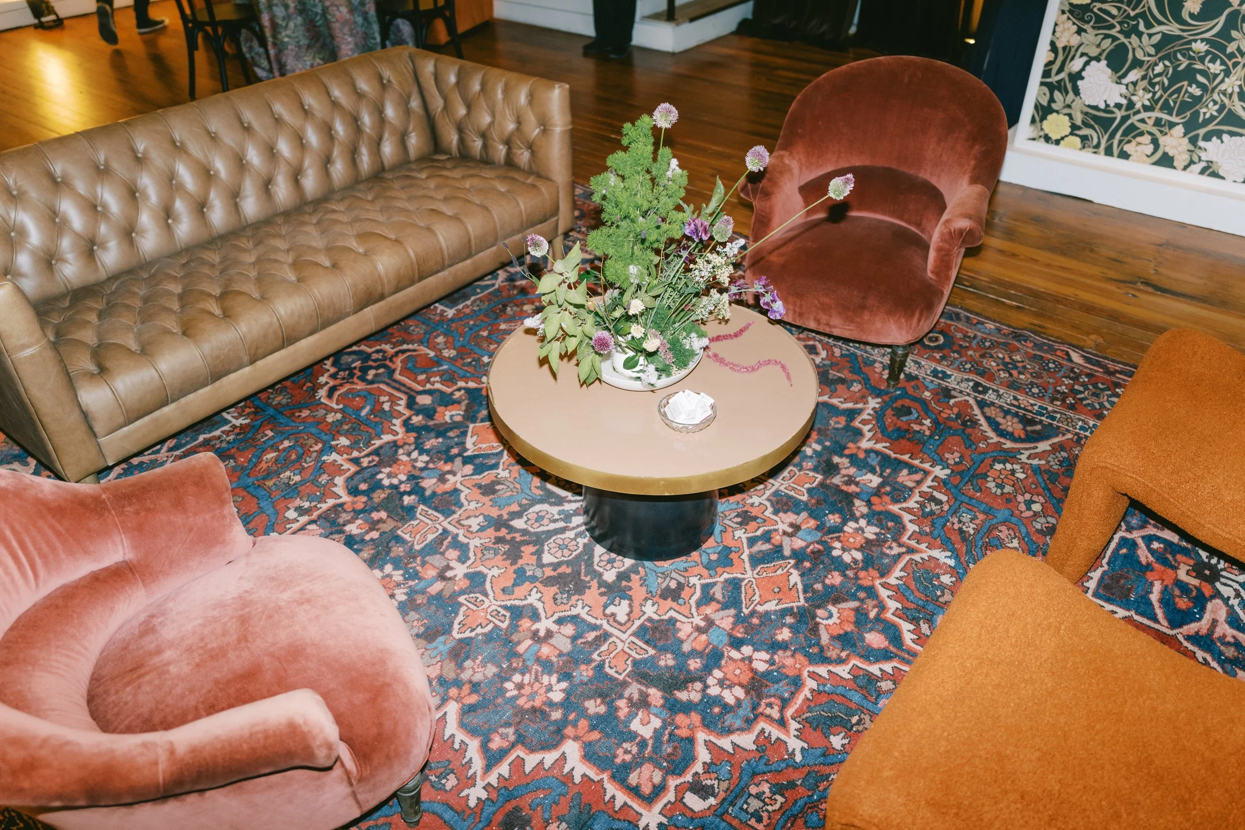 Living room with a brown leather tufted sofa, orange velvet armchair, a rounded pink armchair, and a round coffee table with a floral arrangement. The room features a patterned area rug and wooden flooring.