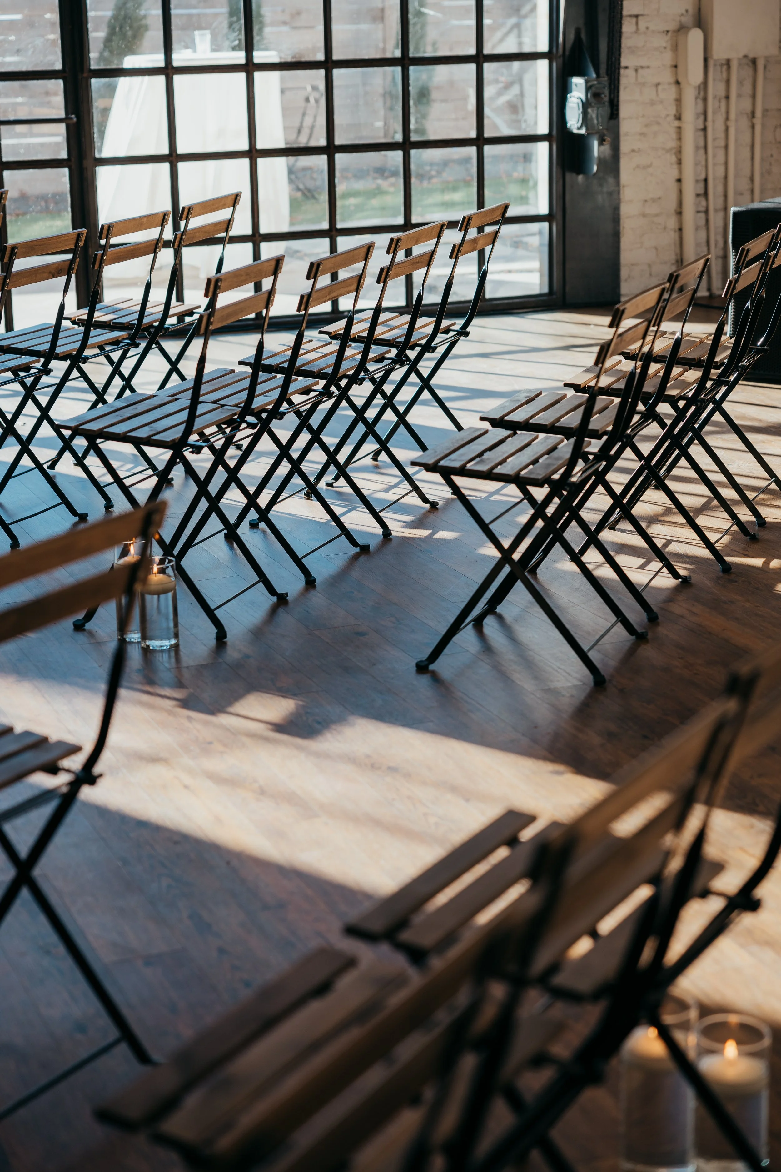 Empty folding chairs arranged in rows inside a bright room with large windows and wooden floors, with sunlight casting shadows.