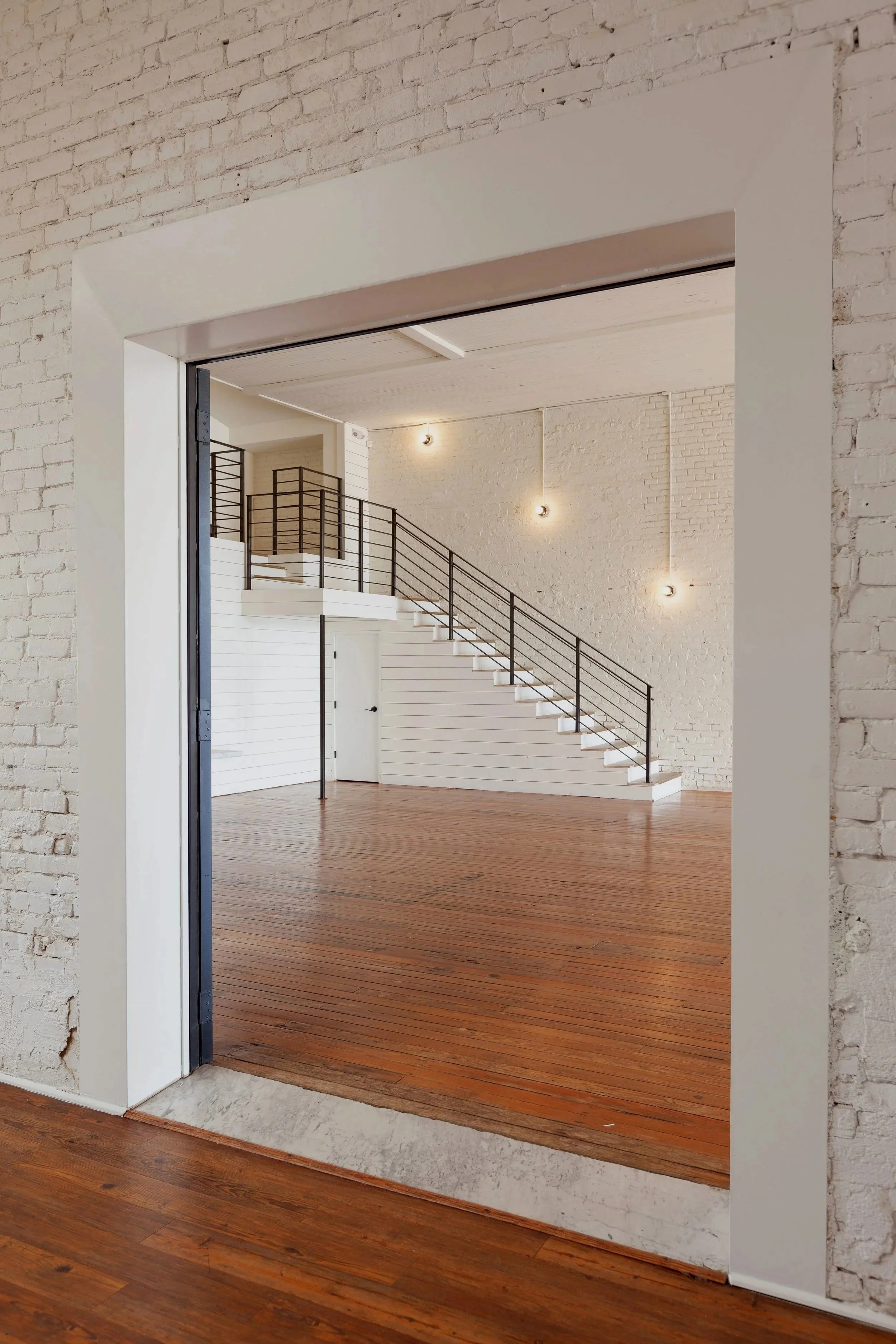 View through a doorway into a spacious, modern, loft-style interior with white brick walls, wooden floors, staircase with black railing, and wall-mounted lights.