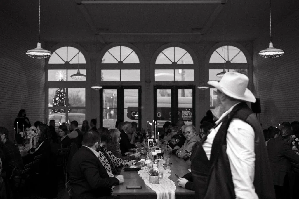 People dining at a long table in a well-lit room with large arched windows and hanging pendant lights, during an indoor event or gathering.