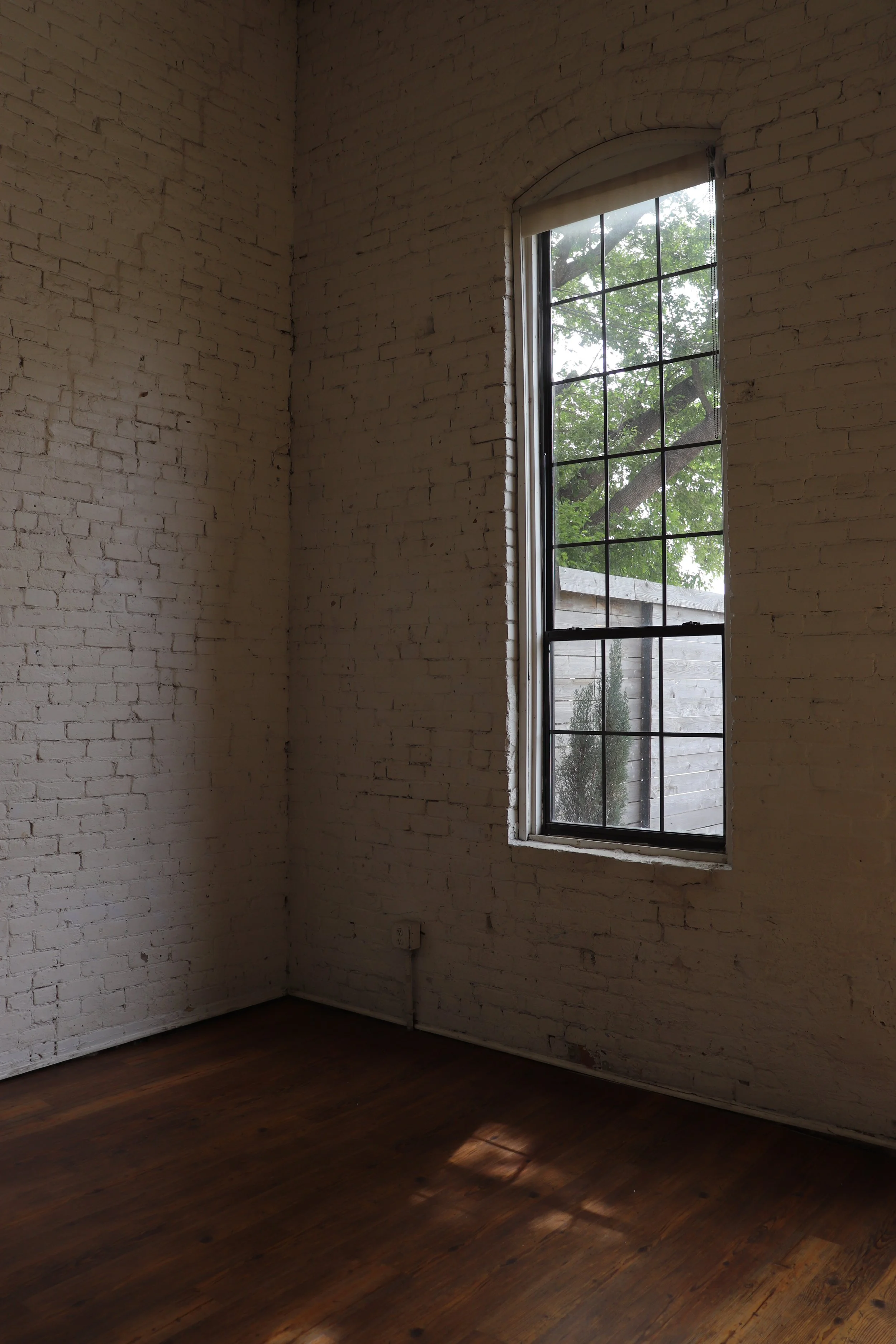 Empty room with white brick walls, a tall window with black trim, and wooden floorboards, with light coming through the window.