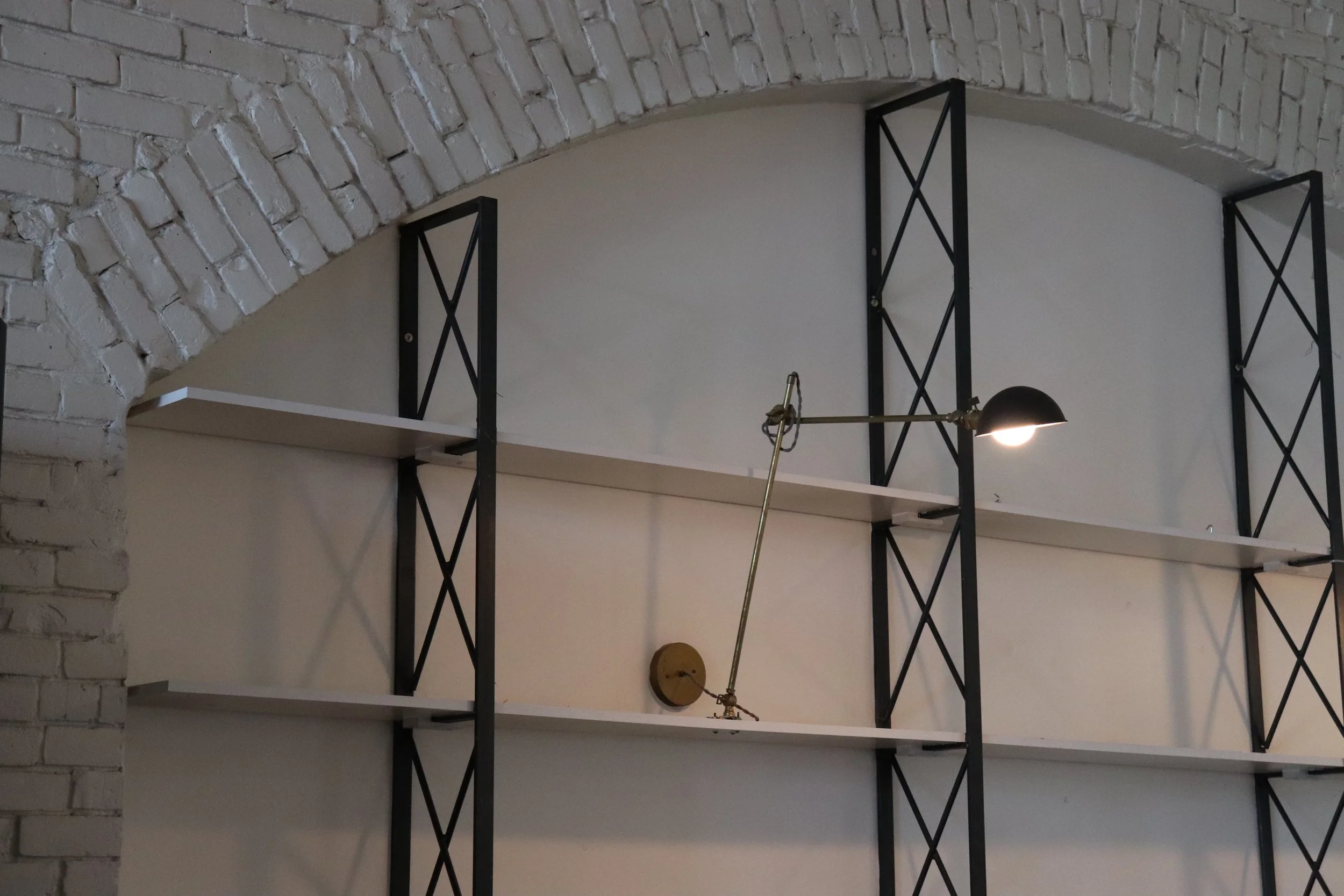 Empty white and black shelving unit mounted on a wall under a brick arch ceiling, with an adjustable wall lamp illuminating the shelves.