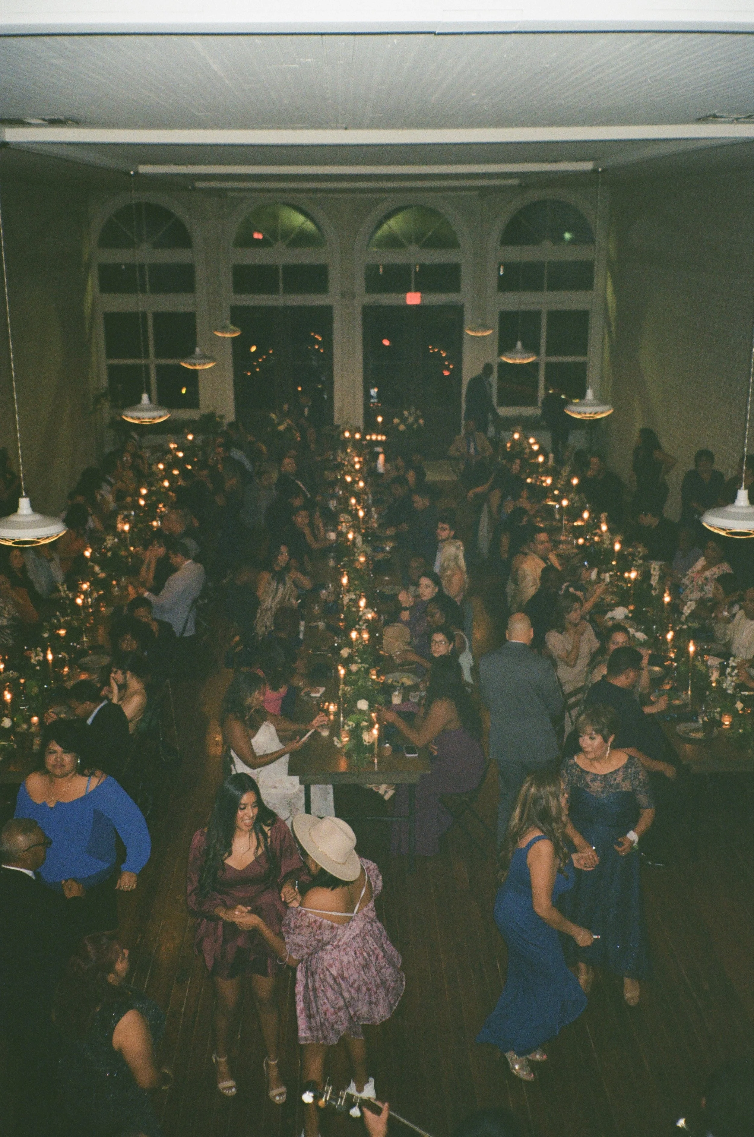 A festive evening dinner event with long tables decorated with candles and greenery, filled with people socializing and dancing in a decorated hall with large arched windows.