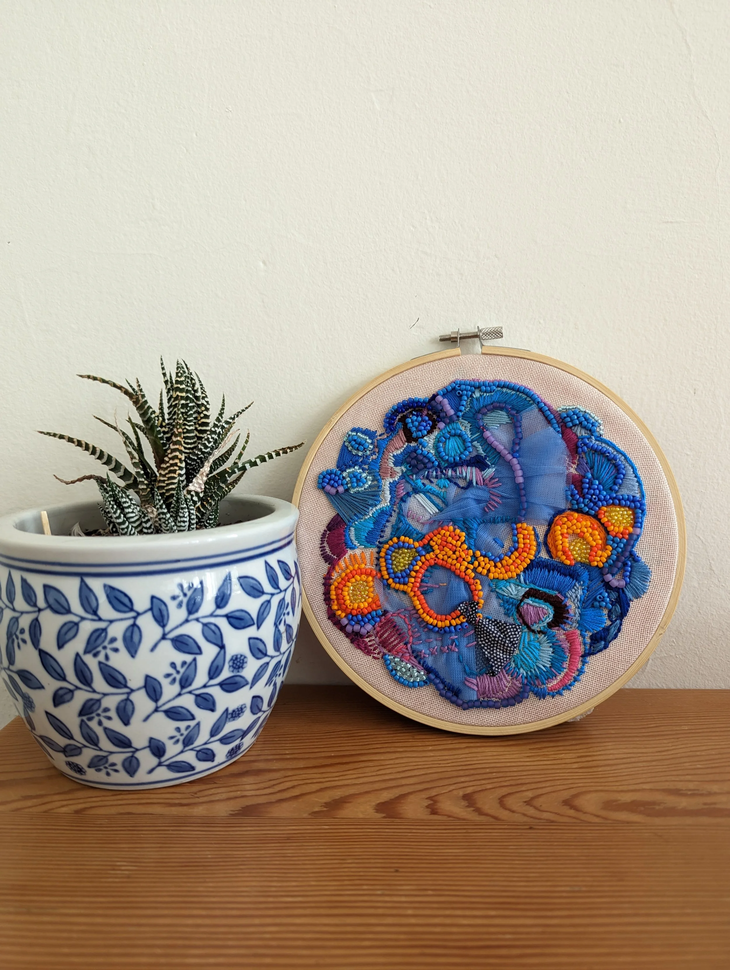 A potted succulent plant with striped green and white leaves on a wooden surface next to a circular embroidery hoop with colorful floral and abstract embroidery on fabric.