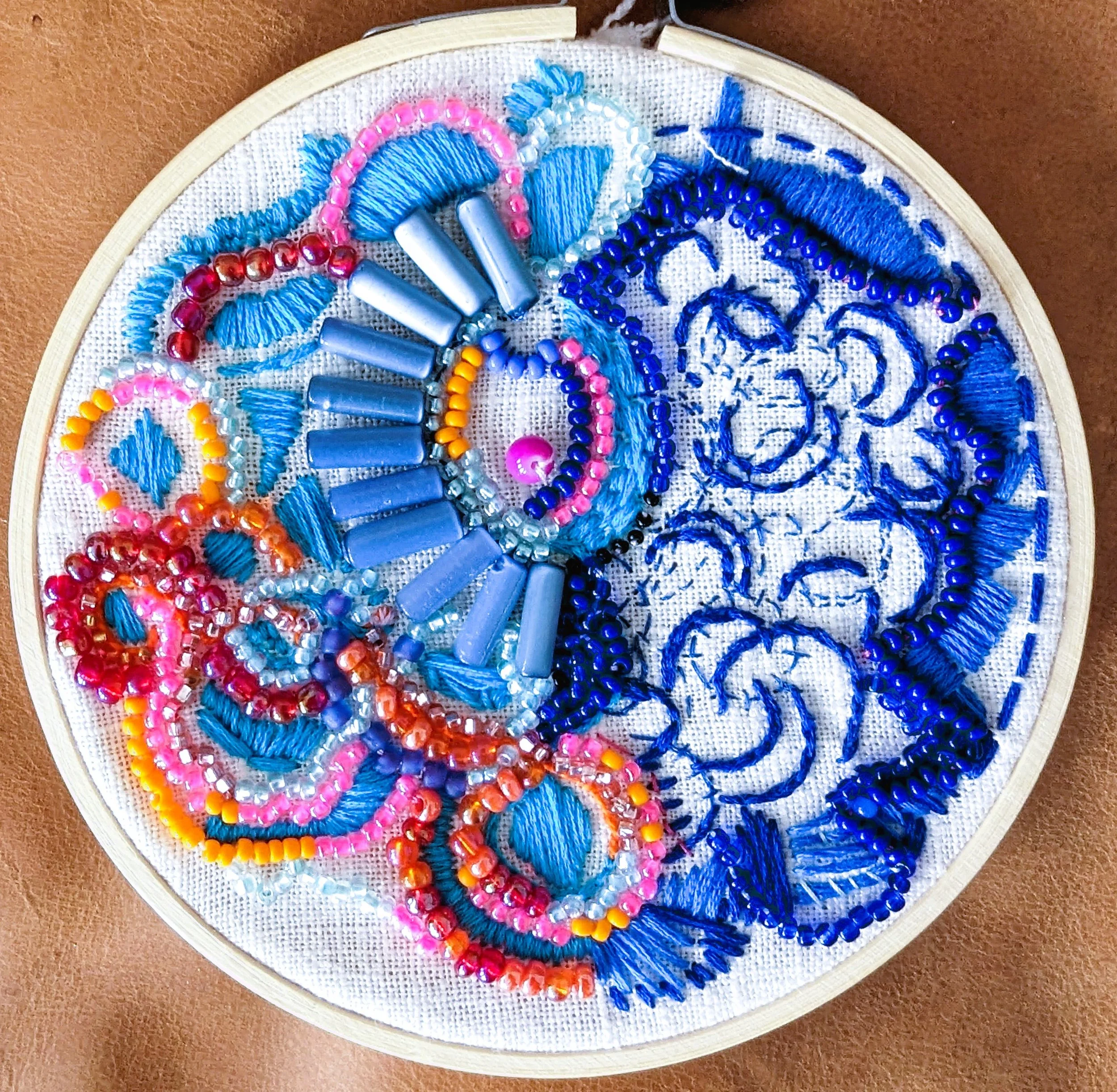 A round embroidery hoop containing a partially stitched colorful beaded and thread embroidery design on white fabric. The design features swirling patterns in shades of blue, pink, orange, and red with various beads and stitched details.