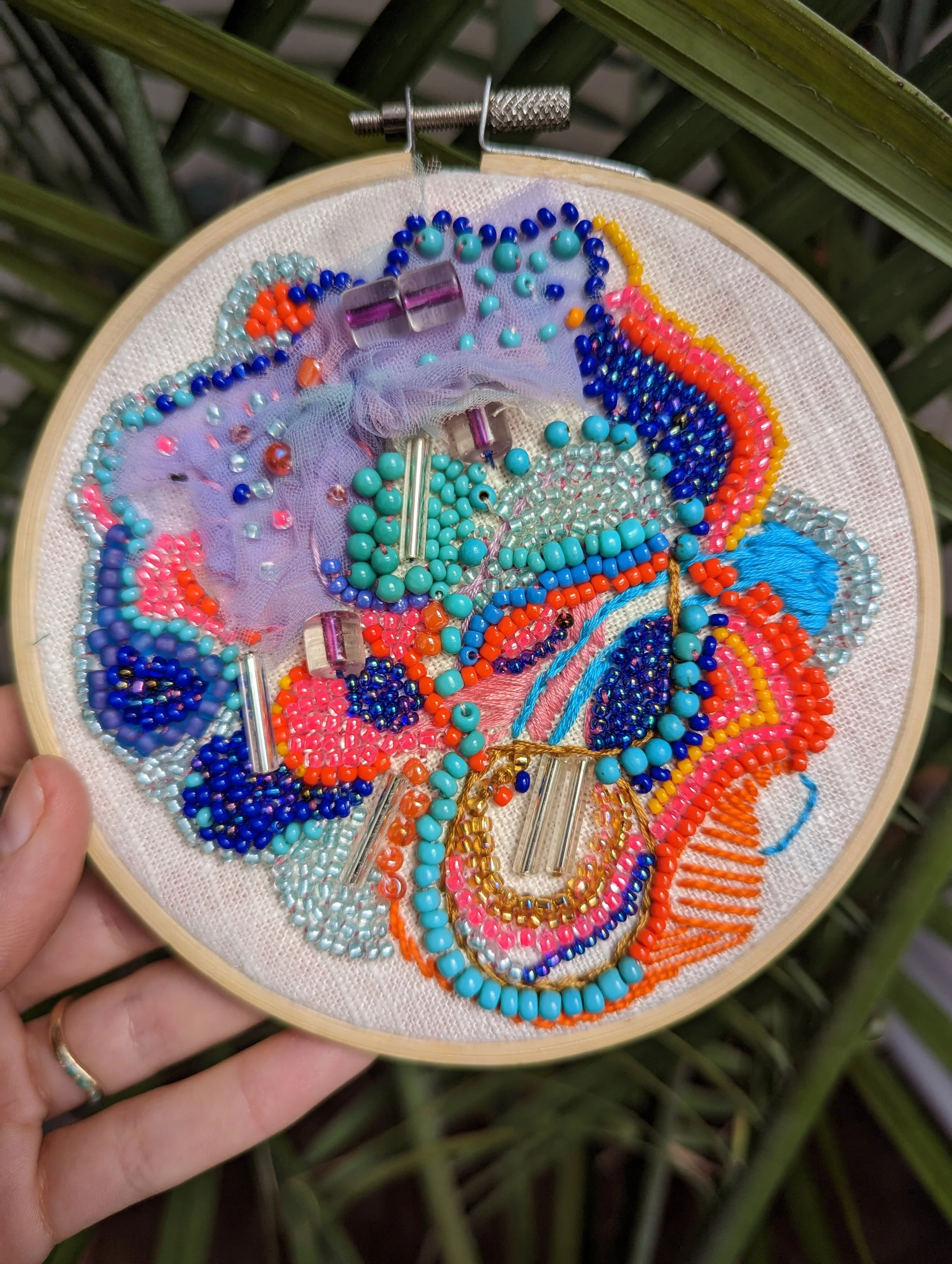 A colorful embroidery hoop featuring various patterns and beads in blue, pink, orange, purple, silver, and gold, with some tulle fabric, held by a hand outdoors among green plants.
