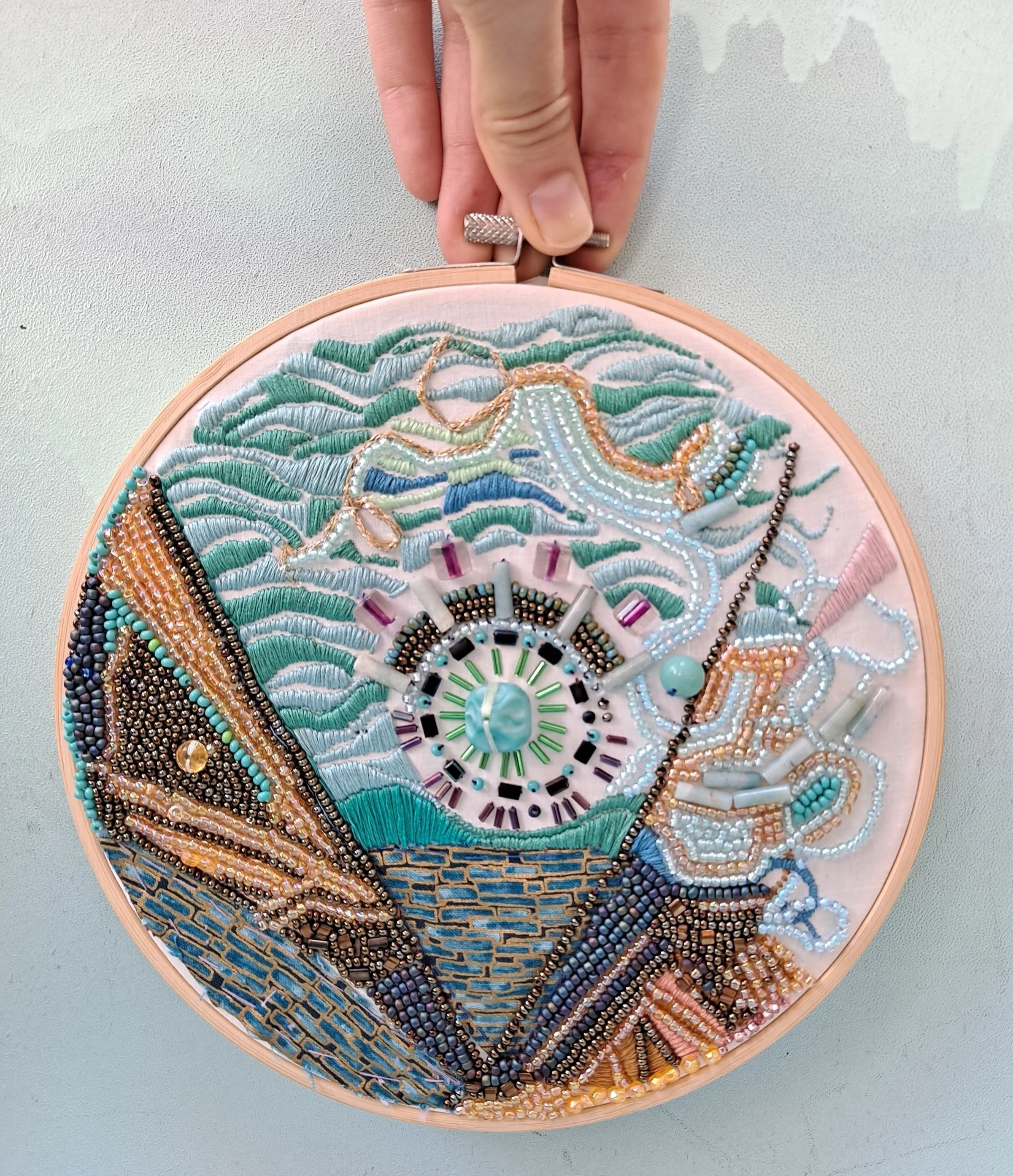A person holds a wooden hoop embroidery hoop, which contains an intricate piece of art made with beads and embroidery threads depicting a still life scene with a purse, a necklace, and some other accessories, all in pastel and metallic colors.