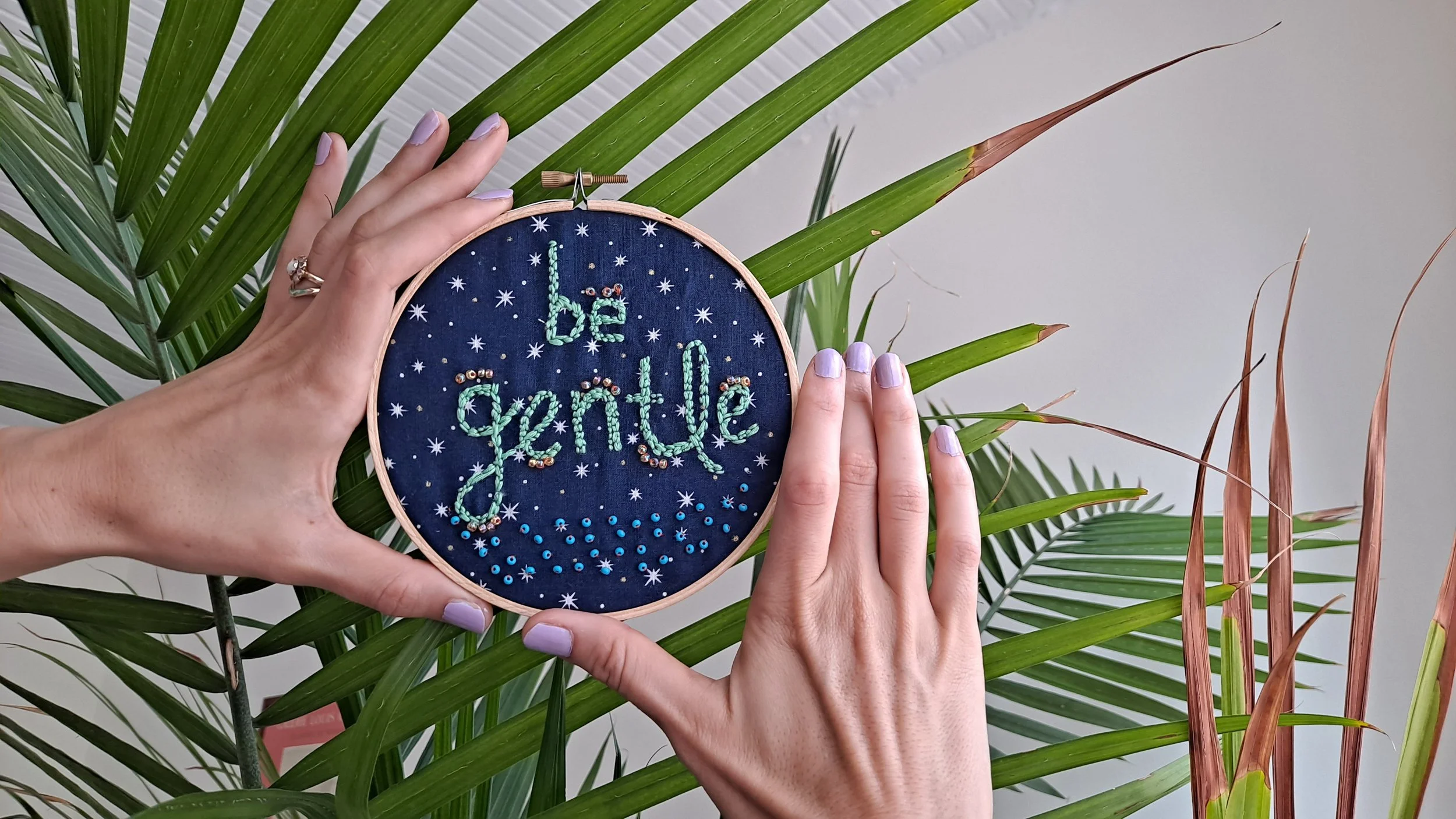 Hands holding an embroidery hoop with the words "it's grateful" stitched on navy fabric, surrounded by star patterns, against a backdrop of green houseplant leaves.