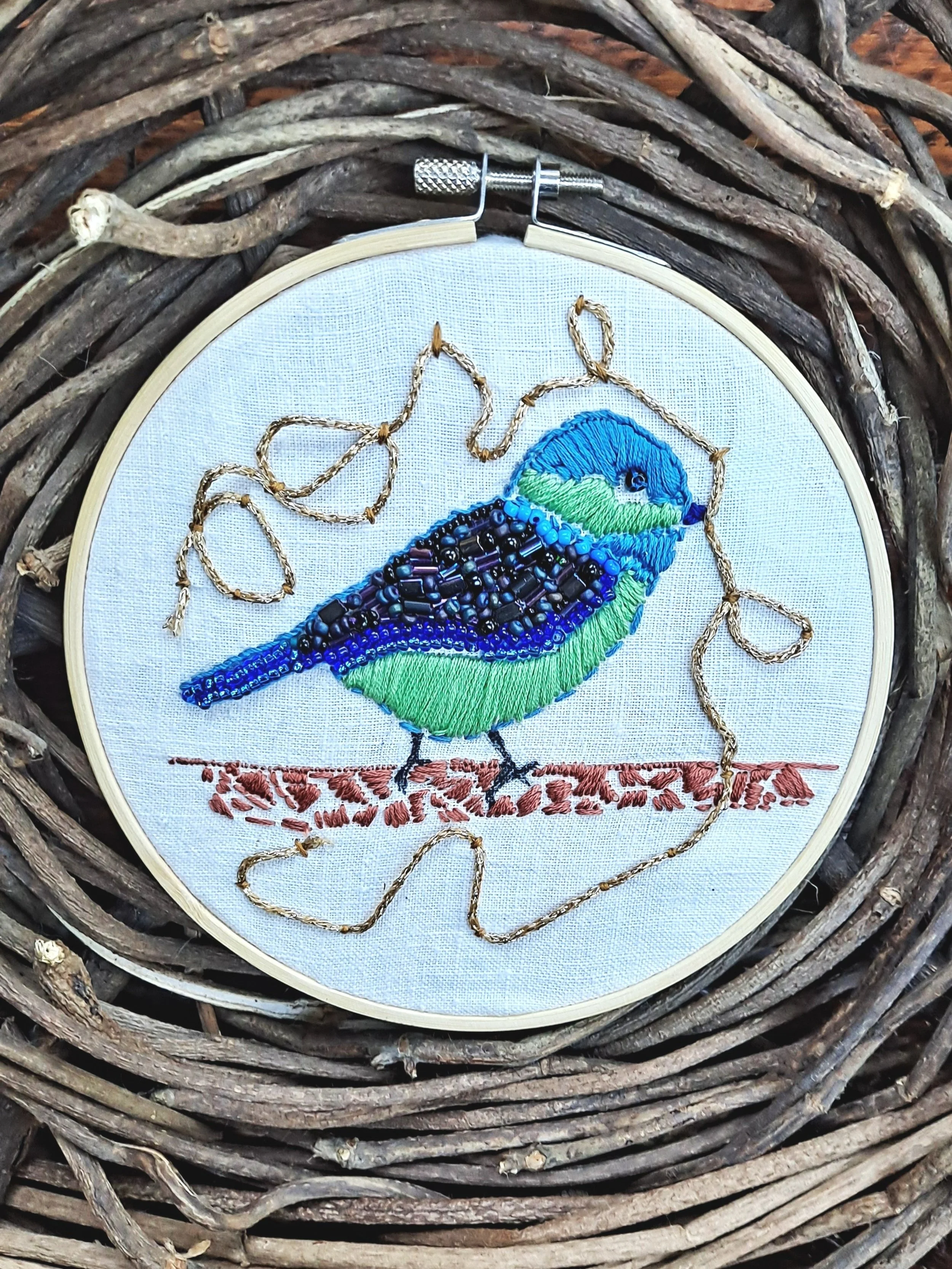 Embroidery of a colorful bird with blue, green, and black feathers, sitting on a branch with a gold thread outline around it, all placed within a wooden embroidery hoop resting on a woven twig background.