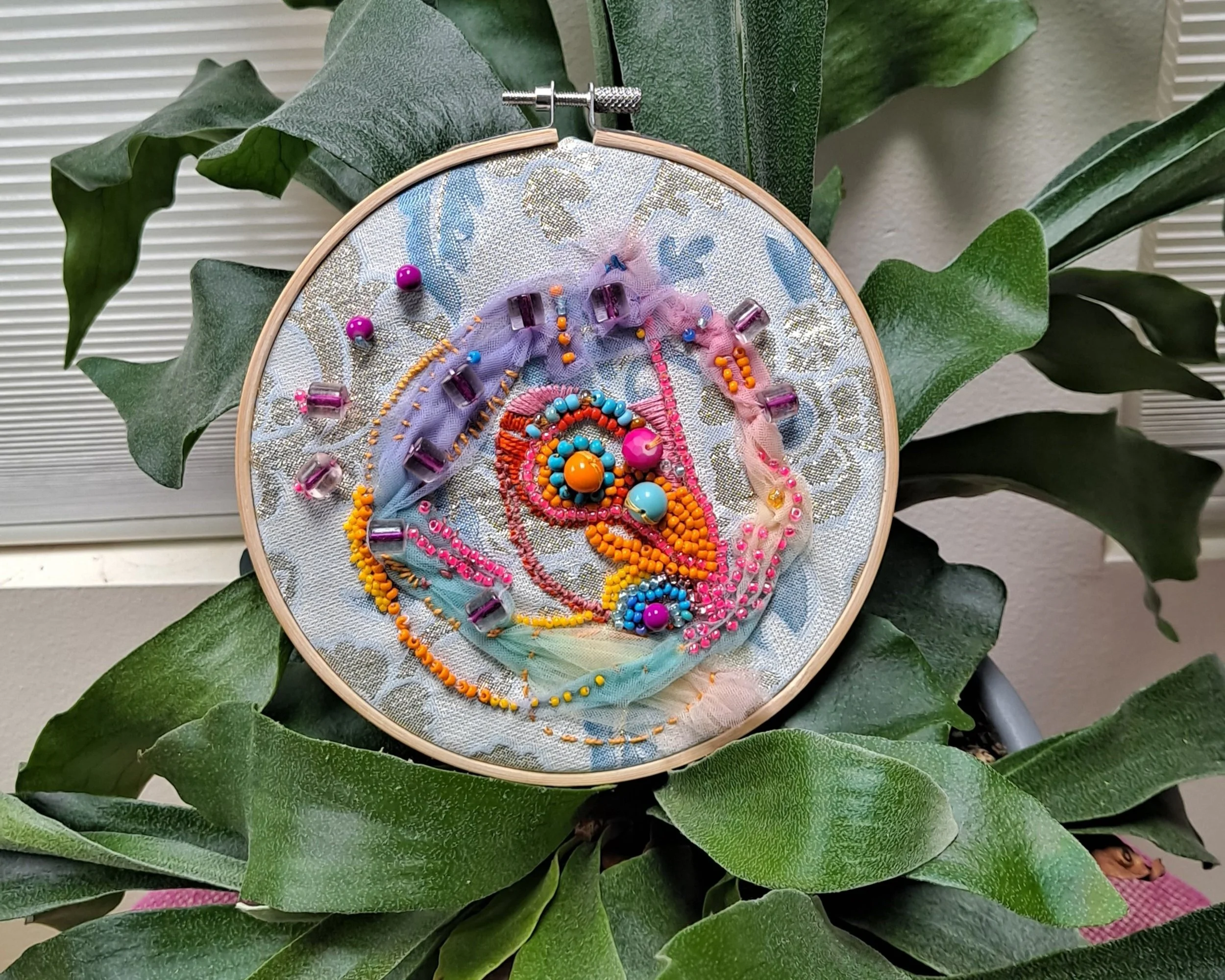 Colorful embroidery hoop art featuring a stylized bird design with bright beads and thread, positioned among green houseplant leaves.