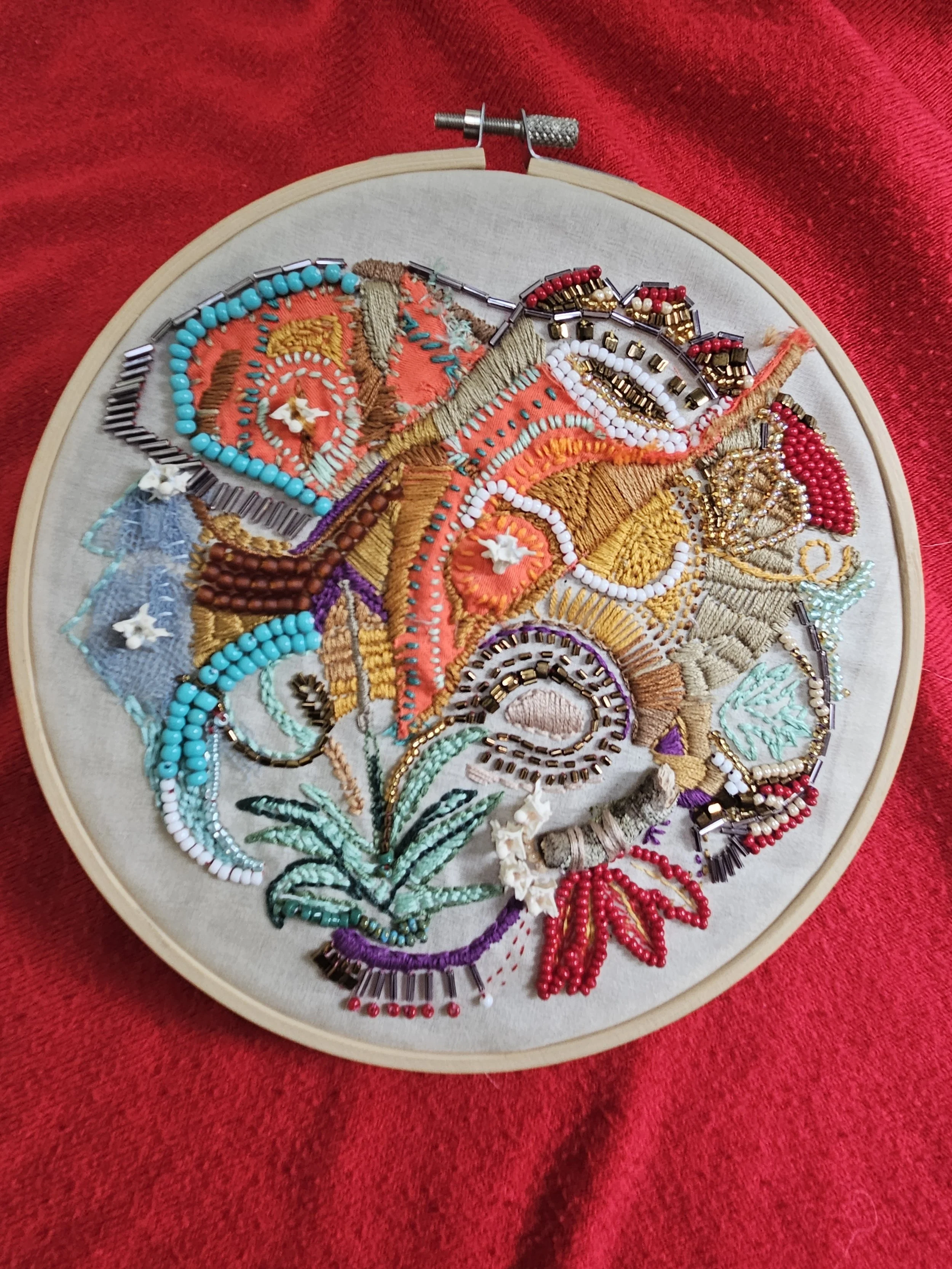 Colorful embroidered textile in a circular shape inside a wooden embroidery hoop, placed on red fabric.