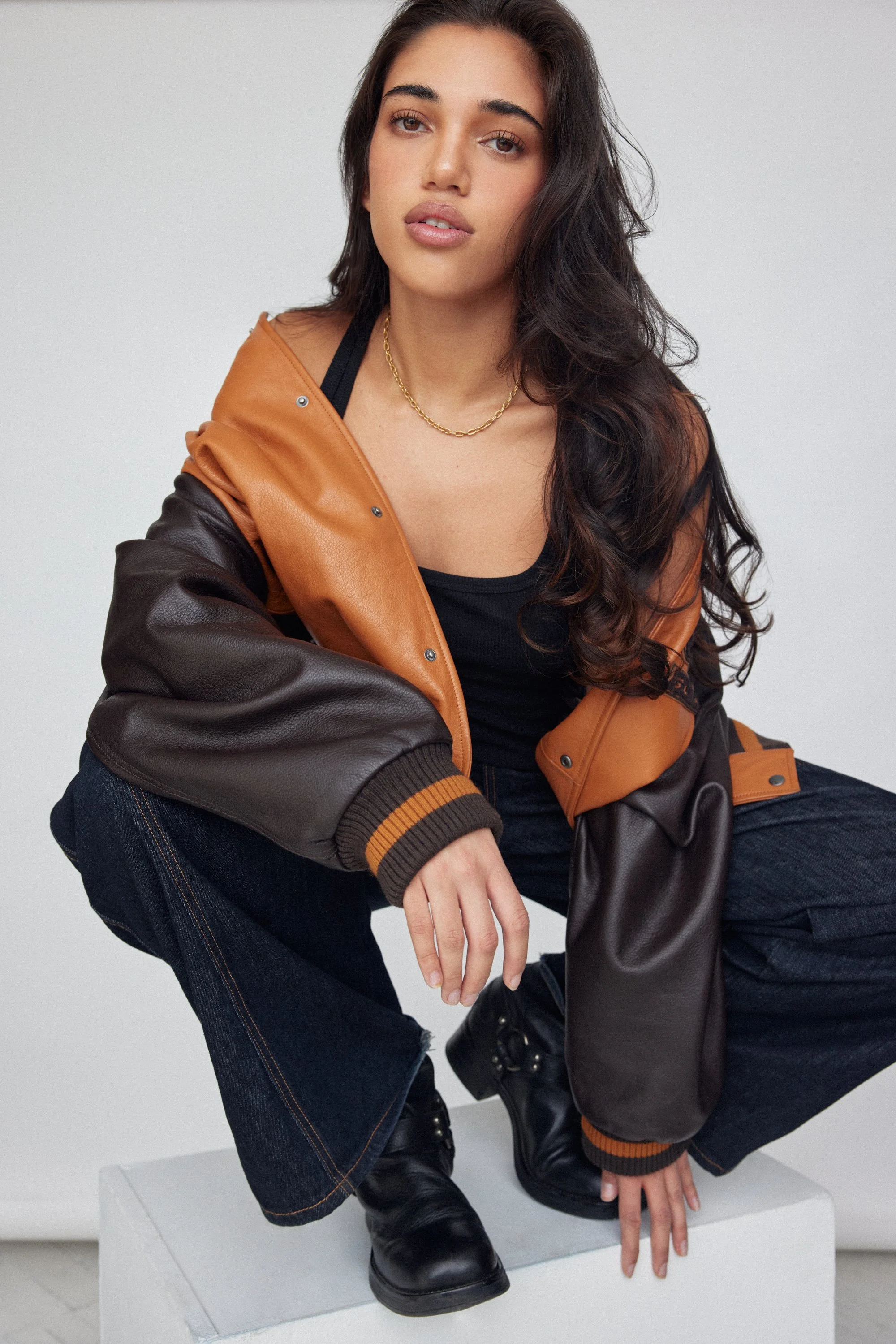 A young woman with long dark hair, wearing a black top, a two-toned brown and tan leather jacket, dark jeans, and black boots, crouching on a white platform against a plain gray background.