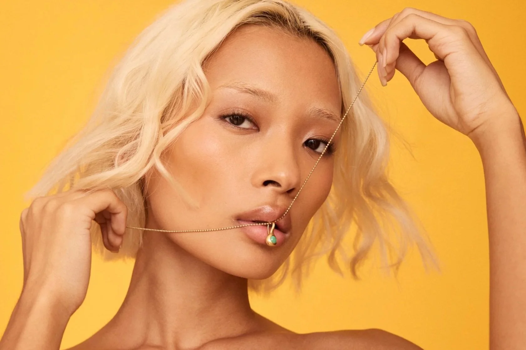 Portrait of a woman with short, wavy blonde hair holding a gold necklace with a turquoise pendant, against a yellow background.